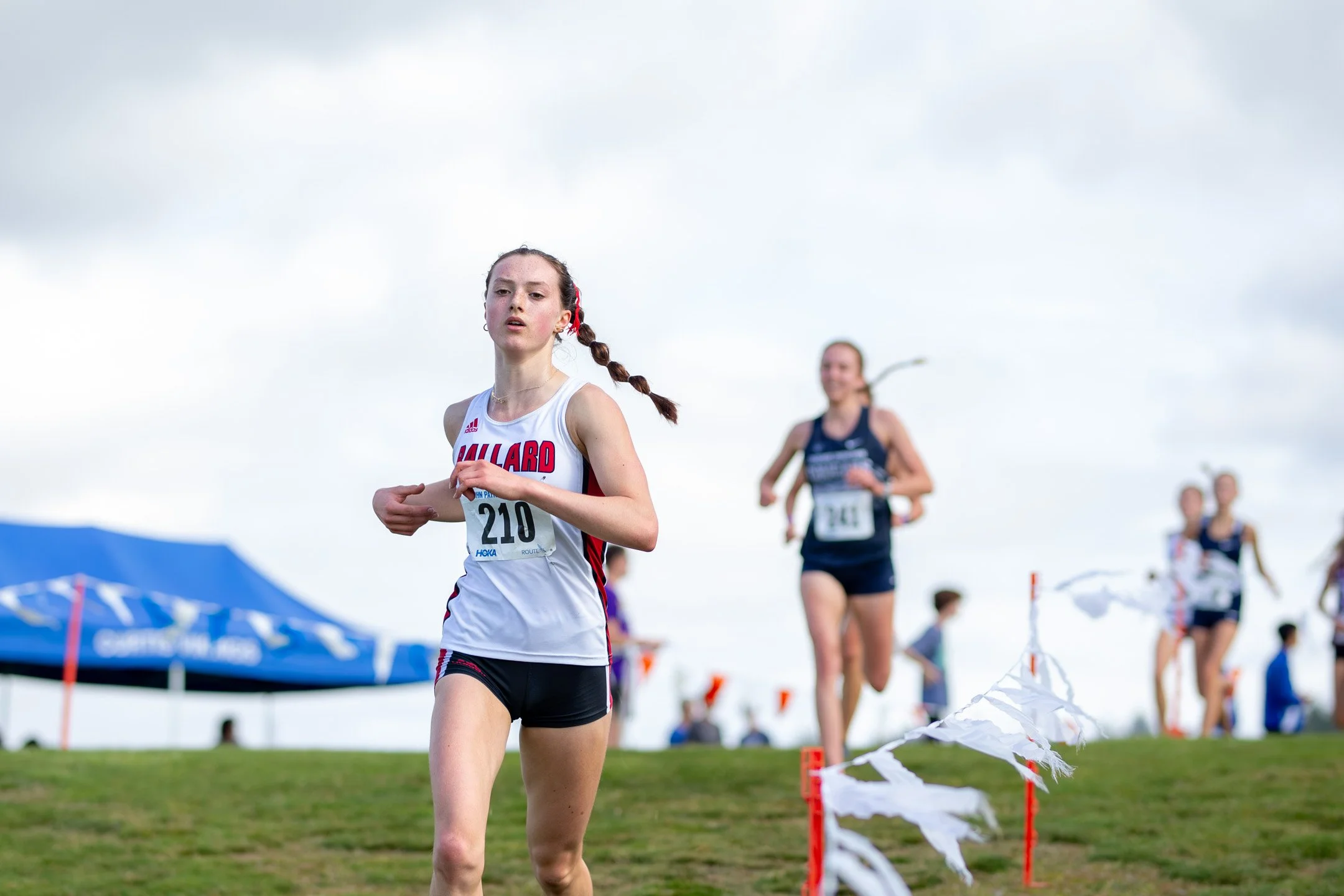 High school girl in a Ballard singlet, bib 210, running alone on grass.