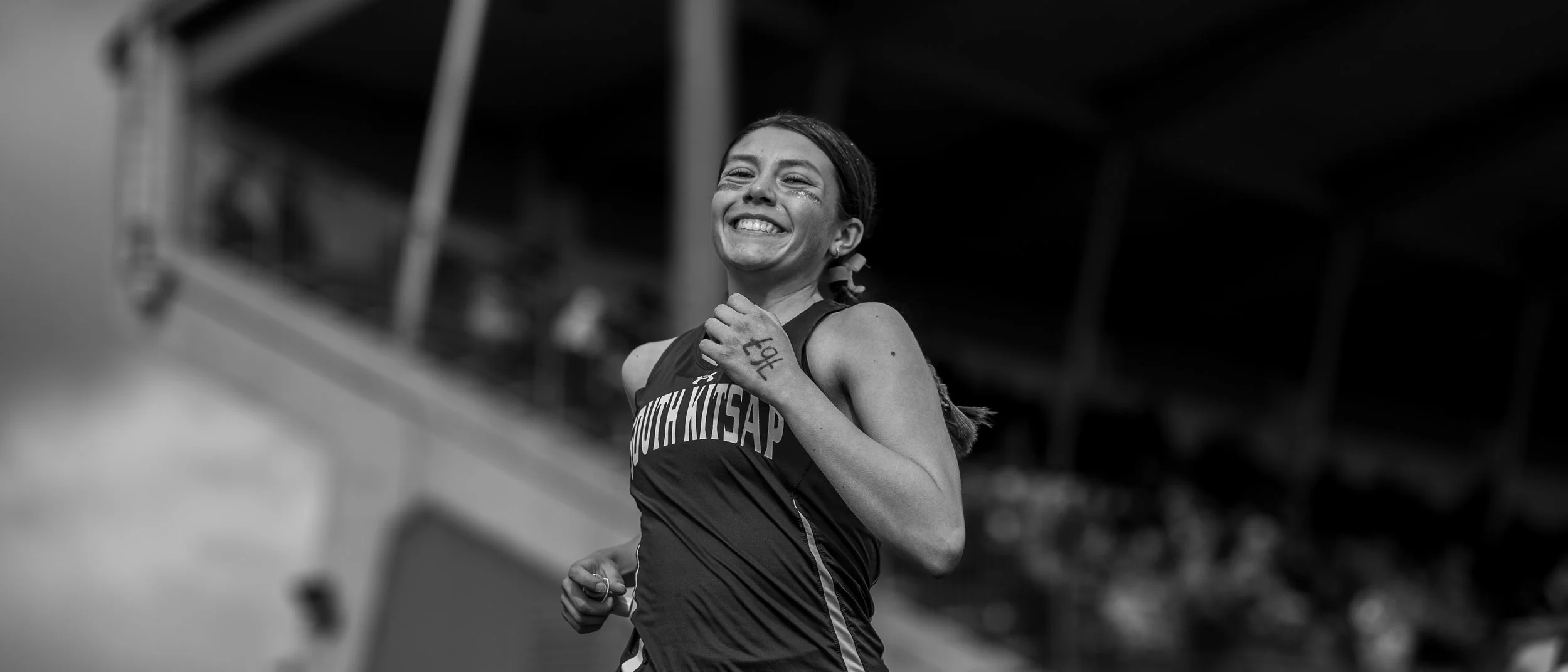 A black and white close-up portrait of a smiling female runner from South Kitsap High School during a track meet at Olympia High School, photographed by Russell Moore.