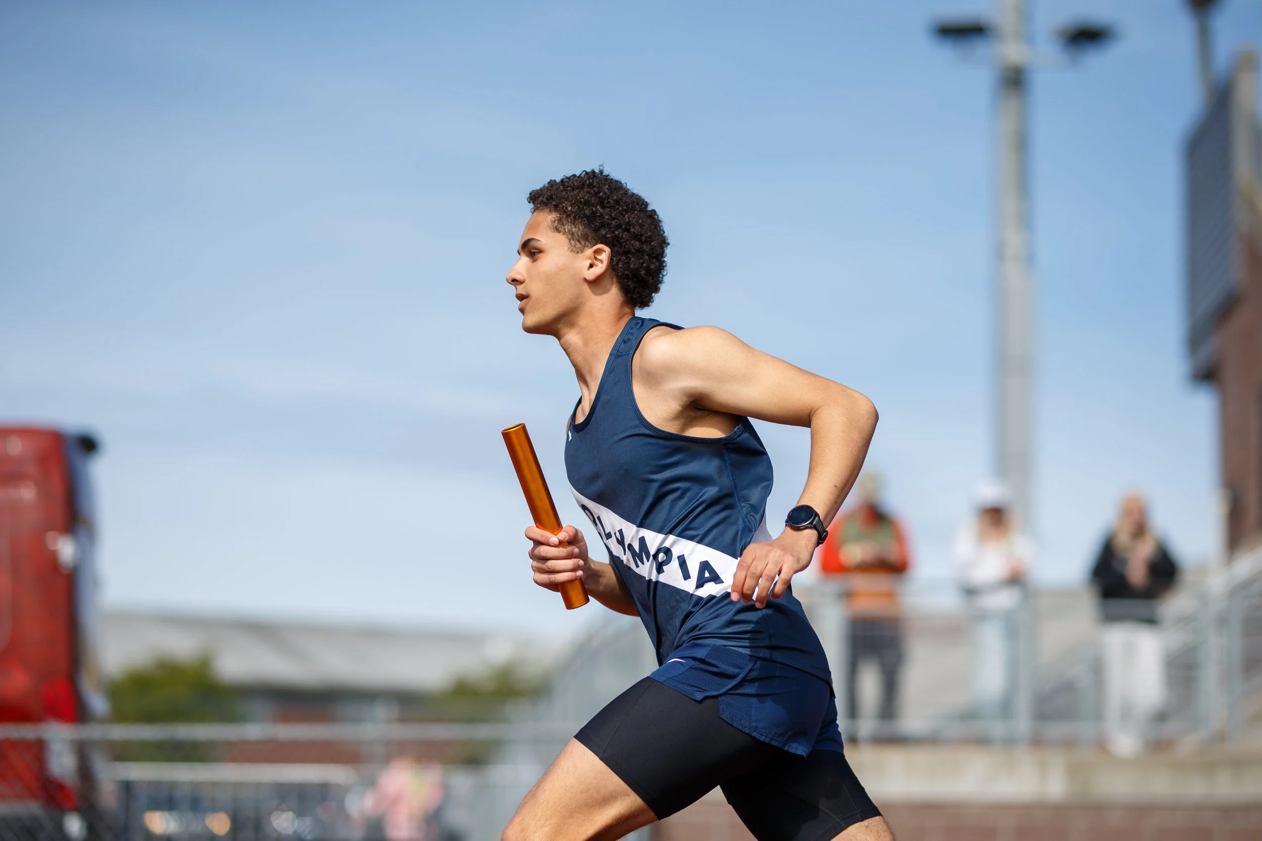 Ayman Ehrlander of Olympia High School running a relay leg, holding a baton, wearing a navy Olympia singlet.