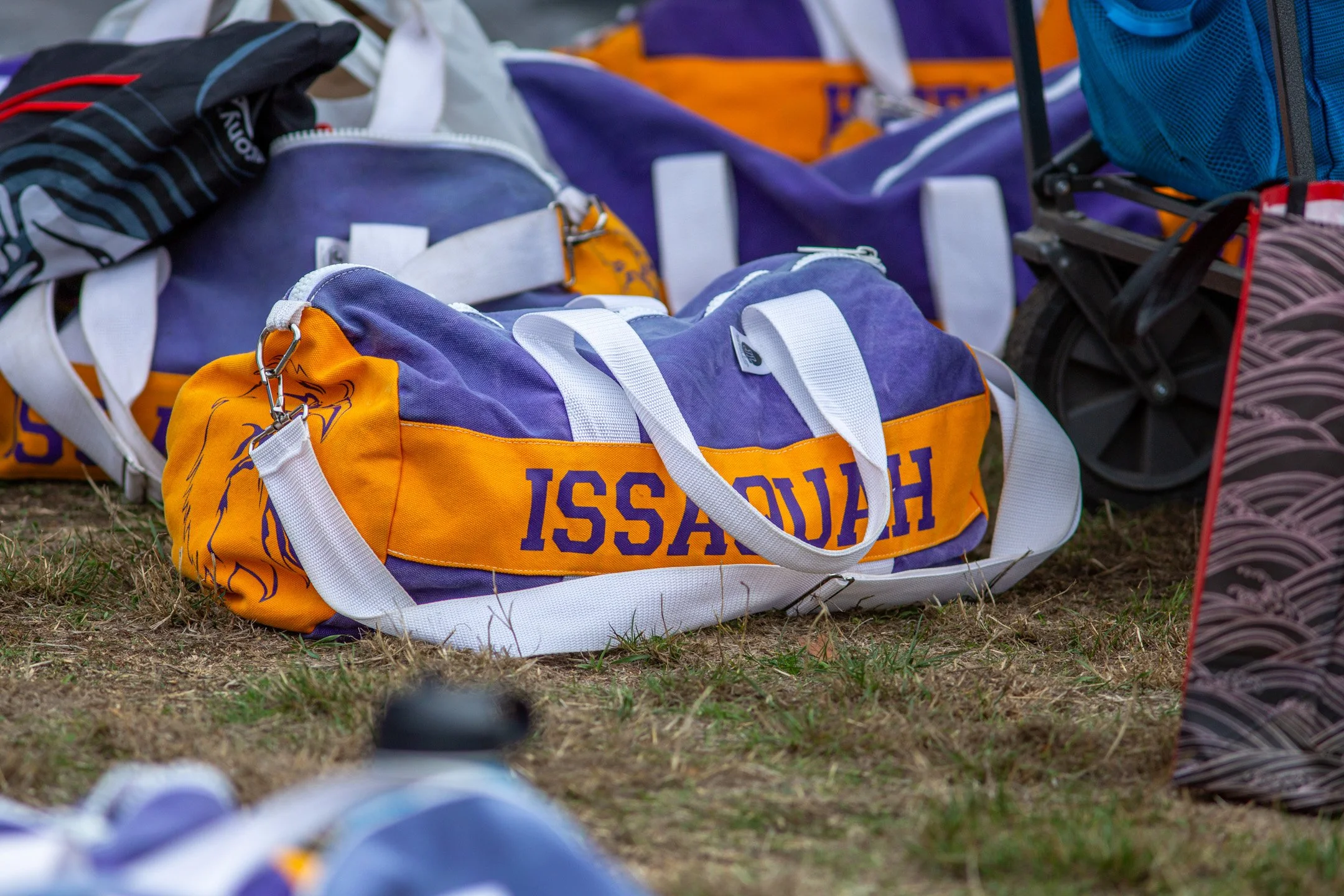 A yellow and blue Issaquah High School duffle bag resting on the grass with other gear under a team tent at a cross country meet.