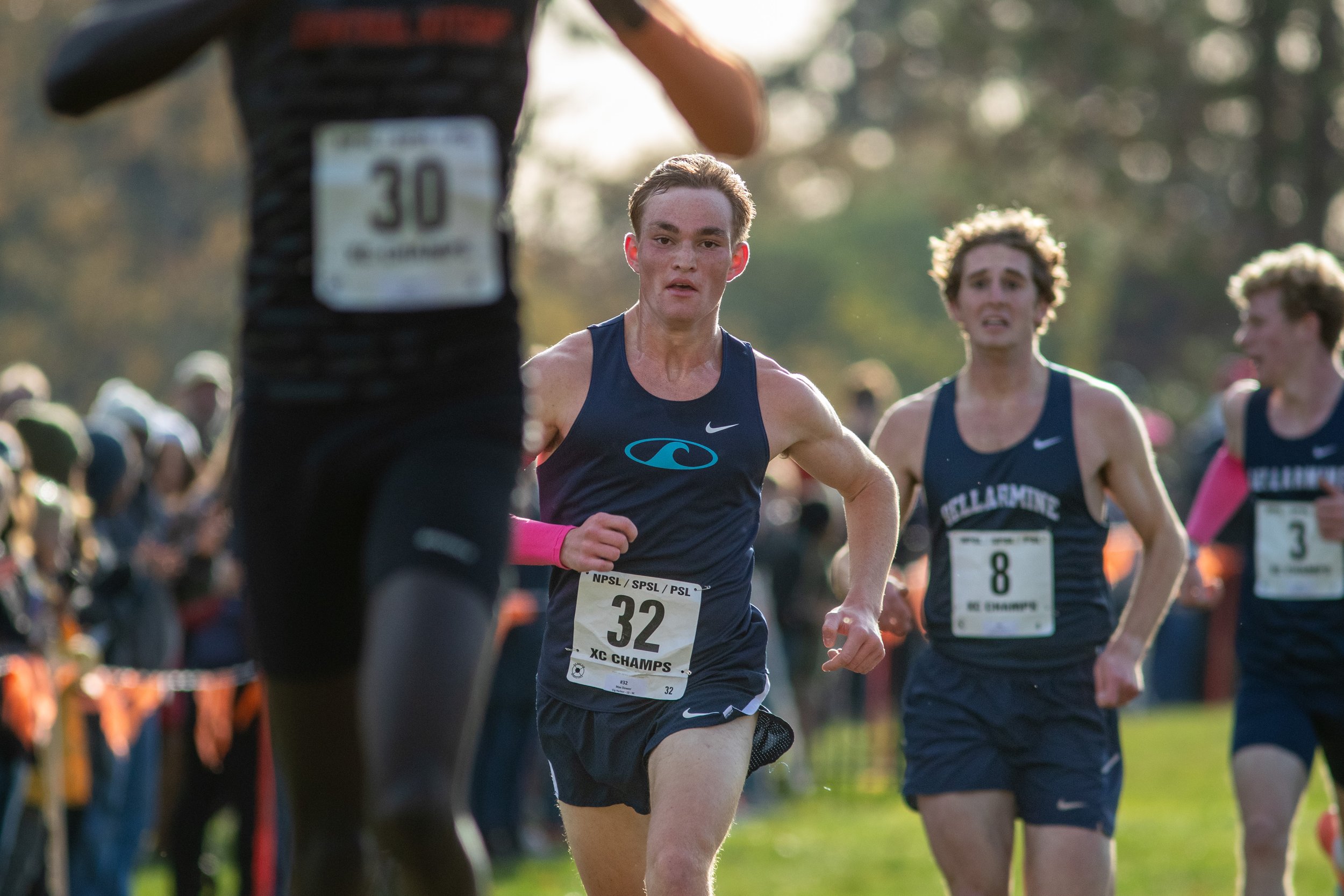 Max Dower of Gig Harbor, bib 32, runs toward the finish in a navy singlet and pink wristband, ahead of two Bellarmine Prep runners in bibs 8 and 3 as a blurred runner passes in the foreground.