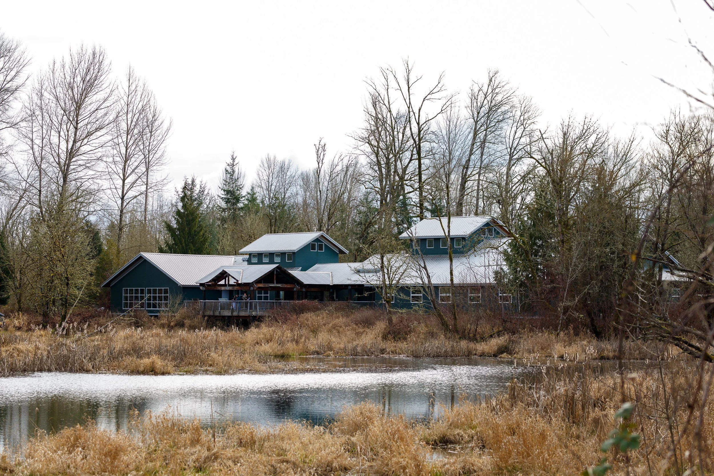 A wide exterior view of the dark green visitor center at Nisqually National Wildlife Refuge in Olympia, WA, framed by dormant winter trees and a small pond, photographed by Russell Moore.