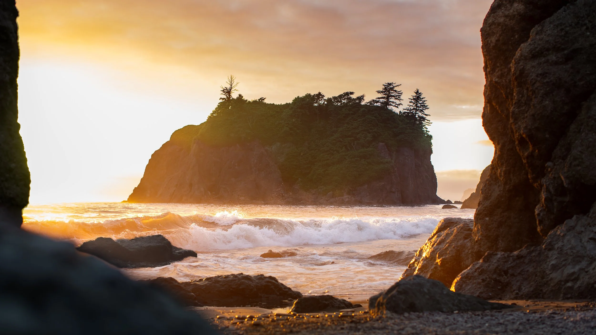 Ruby Beach, WA looking out at Abbey Island at sunset, orange light hitting the water, waves and rocks nearby.