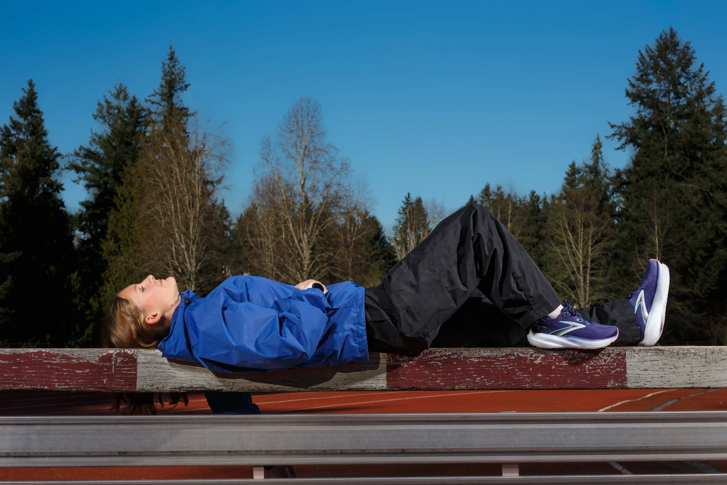 Female athlete in a blue jacket and black pants lying back relaxed on a wooden steeplechase barrier on a red running track with trees and blue sky in the background.