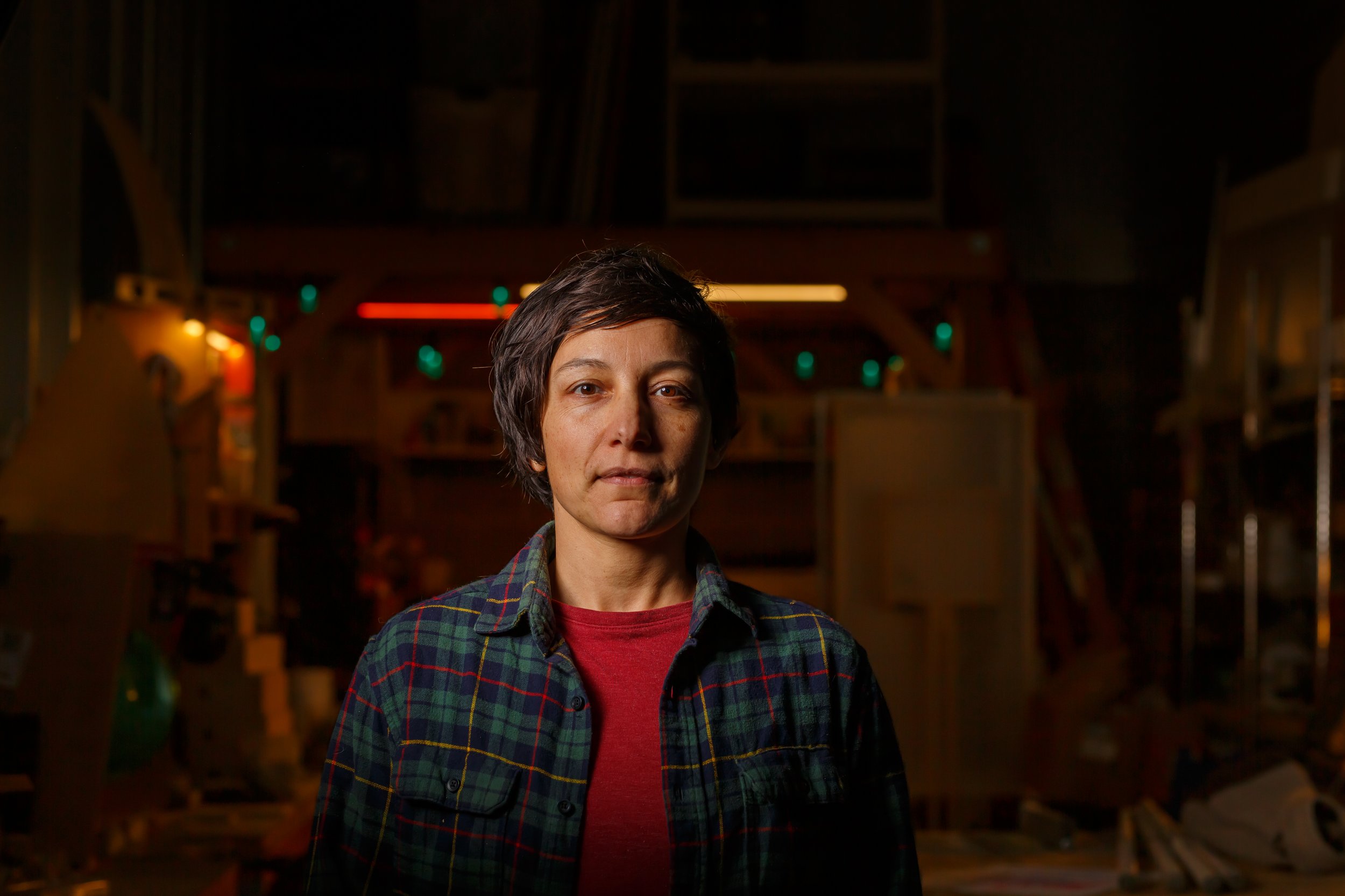 Local non-binary artist Natalie Cobletz stands confidently inside their dimly lit creative workshop in Olympia Washington wearing a plaid shirt and looking directly into the lens.