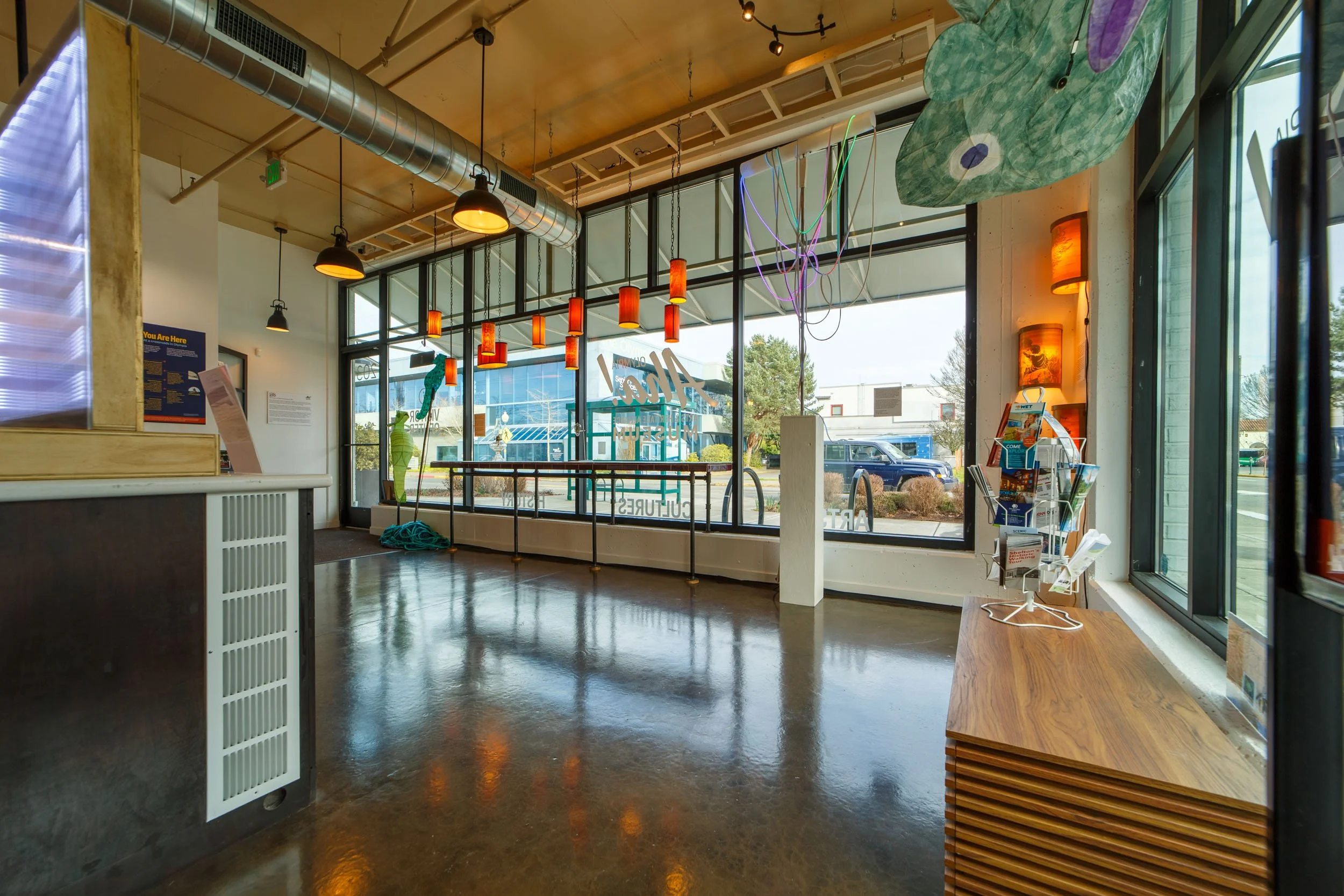 Bright interior architectural photography of a commercial museum lobby in Olympia, Washington, featuring polished concrete floors, exposed ductwork, large windows, and decorative hanging pendant lights.