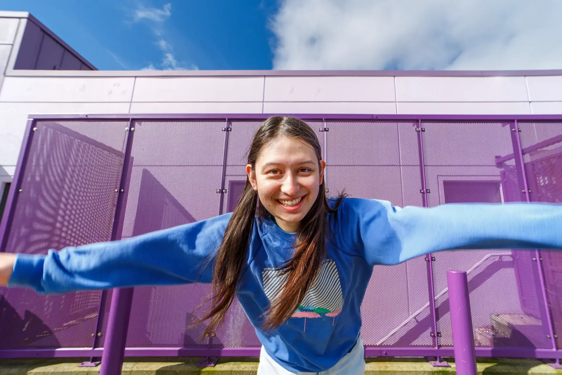 Comedian Nat Penrose leaning aggressively toward the camera lens with arms spread wide, laughing genuinely in front of a bright purple architectural wall under a blue sky.
