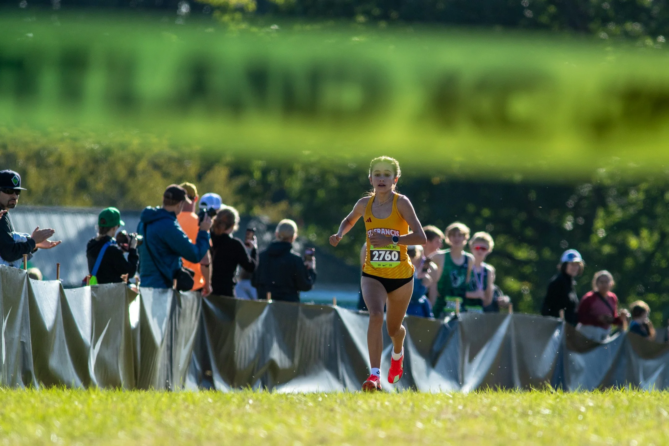 St. Francis sophomore Alyssa Gutierrez in a yellow singlet runs toward the camera, spectators behind her, banner blurred above.