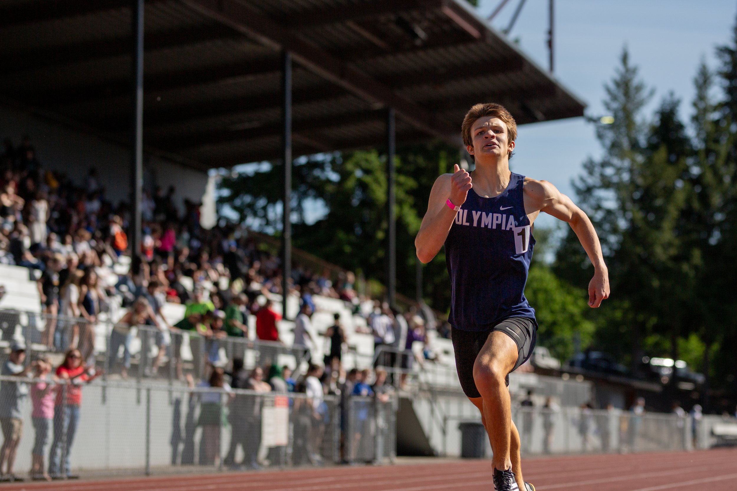 Wesley Stevick of Olympia High School running the track in a navy Olympia singlet, with the stadium crowd in the background.