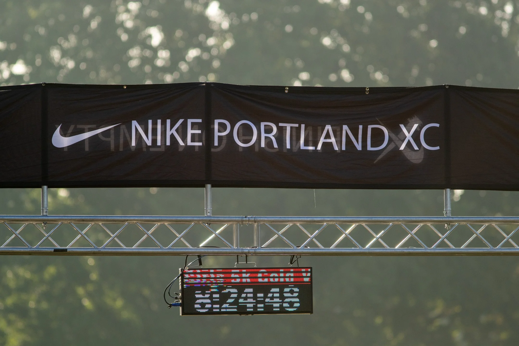 Black Nike Portland XC banner on a metal truss above the finish-line clock, with trees softly blurred behind.