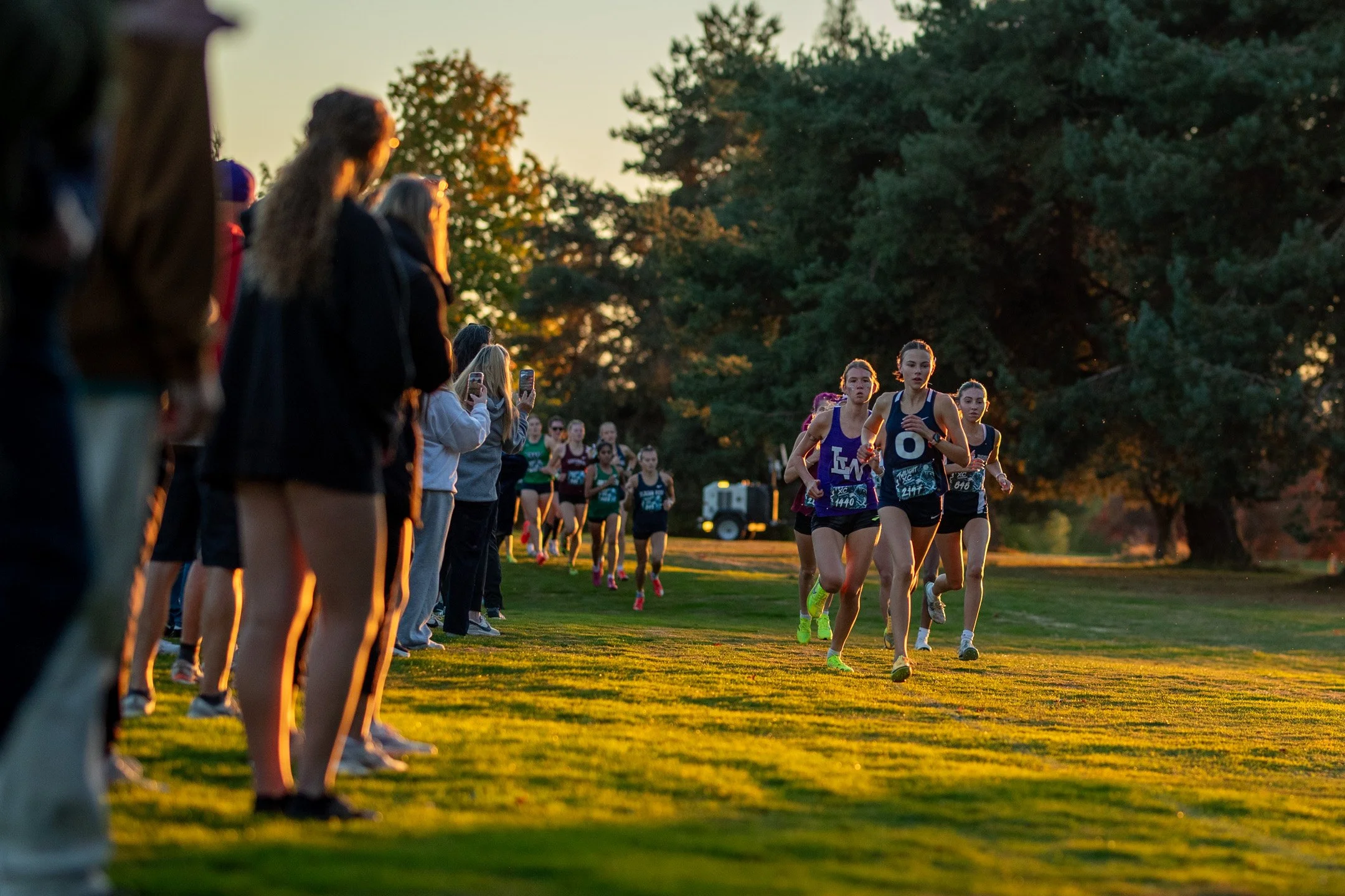 A group of female cross country runners led by Leah Hennen and Elin Beeler running across a golf course in warm evening light past spectators.