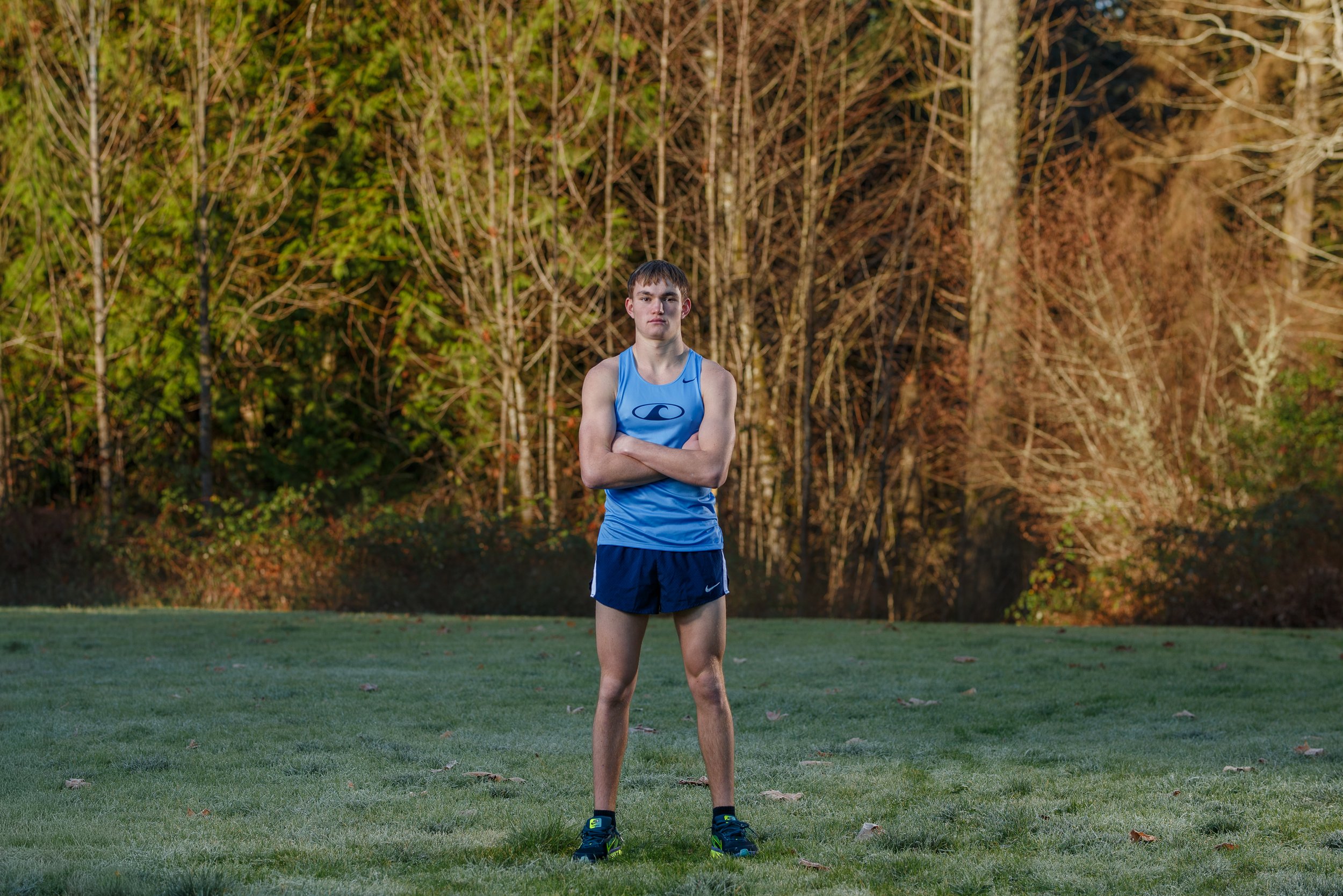 Full body environmental portrait of a male runner standing in a frosty grass field at Sehmel Homestead Park in Gig Harbor, wearing a blue singlet and shorts with morning sun hitting the trees behind him.