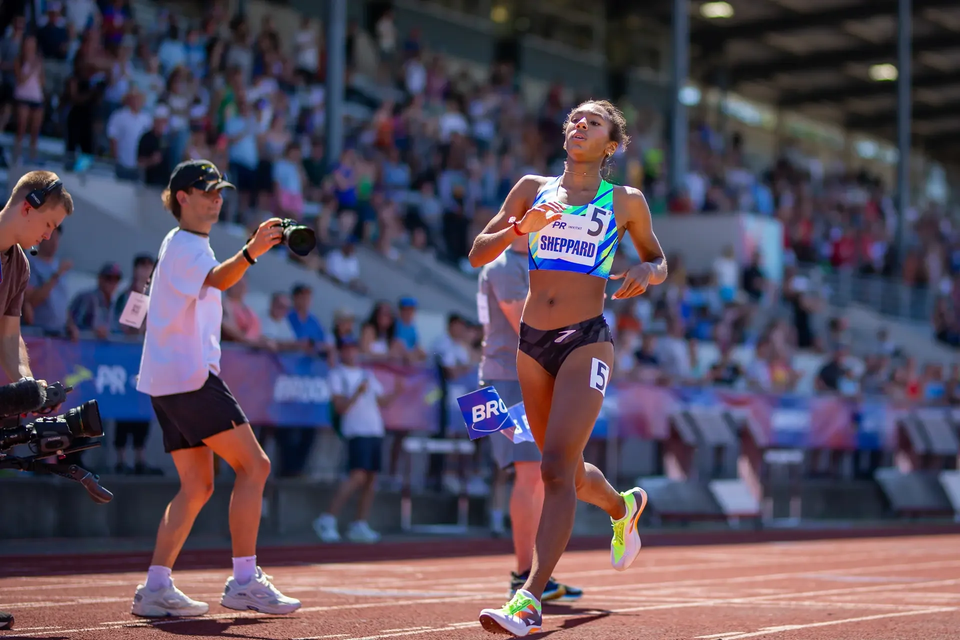 Paige Sheppard of Union Catholic running on the track in a blue and green singlet during her record-breaking 800m race at the Brooks PR Invitational, with photographers and grandstands in the background.