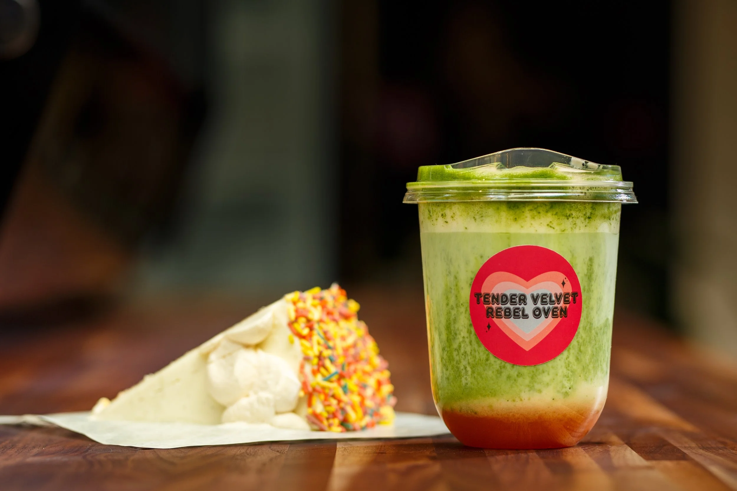 A brightly layered pink and green guava matcha iced drink sits next to a slice of thick white cake covered in colorful sprinkles on a wooden bakery counter at Tender Velvet Rebel Oven.