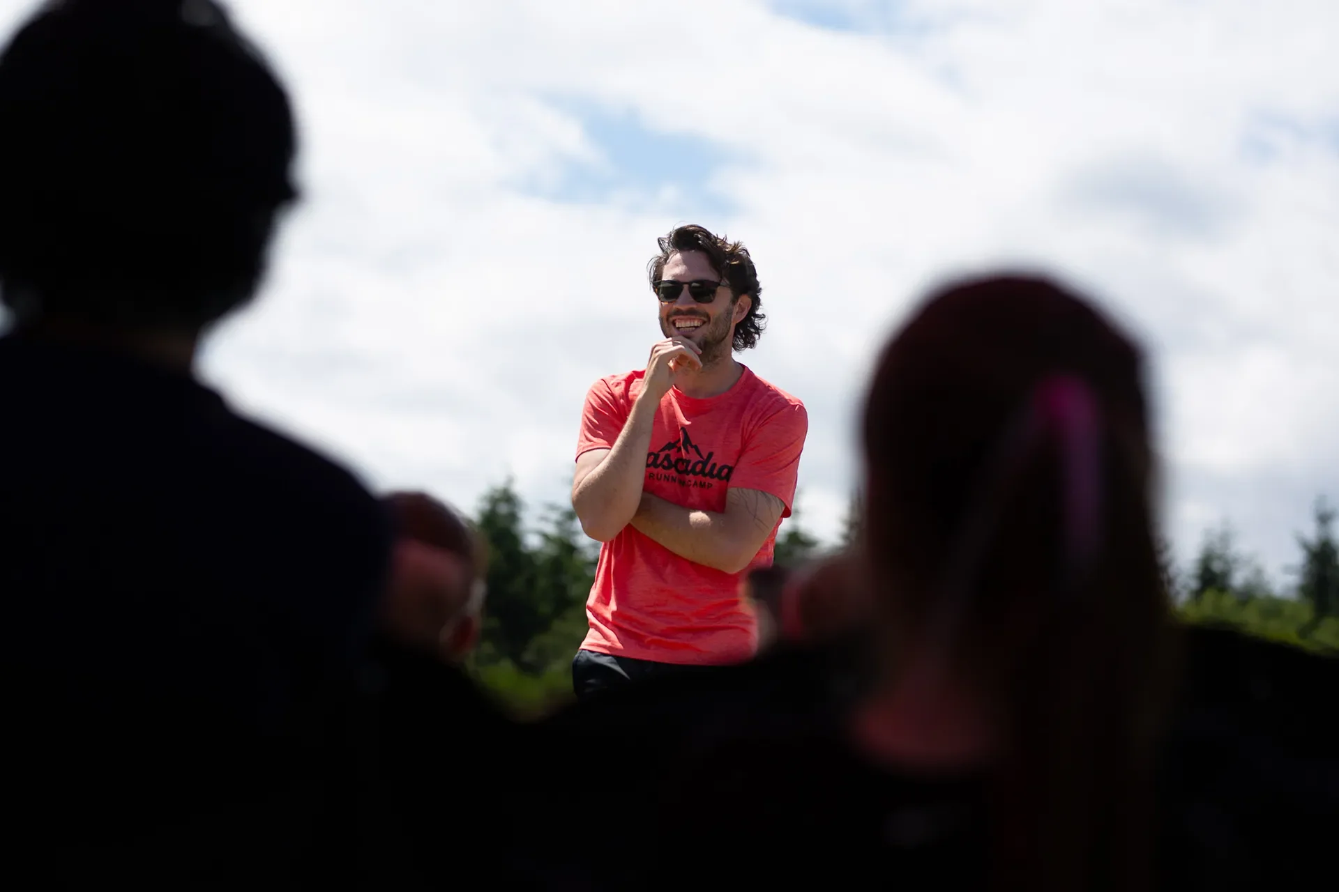 Coach Drew Windle wearing a red Cascadia Running Camp shirt and sunglasses, smiling while talking to a crowd of athletes viewed from behind.