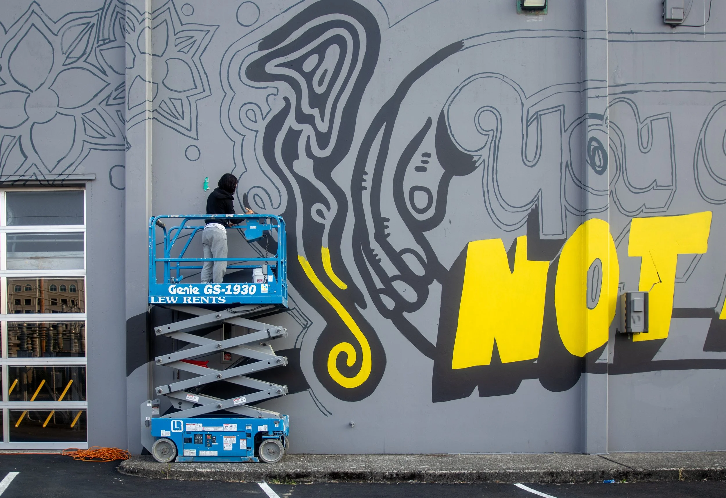 Artist Elisa Del Giudice standing on a blue scissor lift, painting the black outlines of the "You Are Not Alone" mural in Olympia, WA.