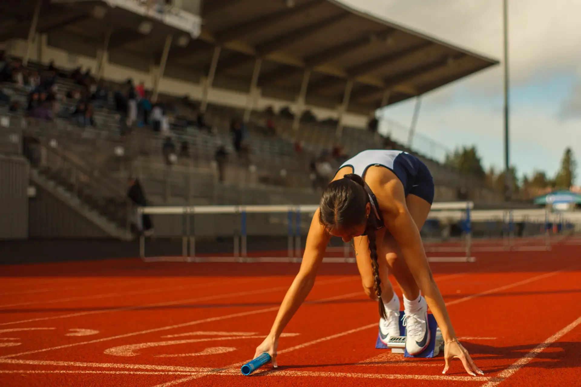 A female track athlete in a blue and white uniform crouched in metal starting blocks holding a blue relay baton on a red all-weather track at Olympia High School, photographed by Russell Moore.
