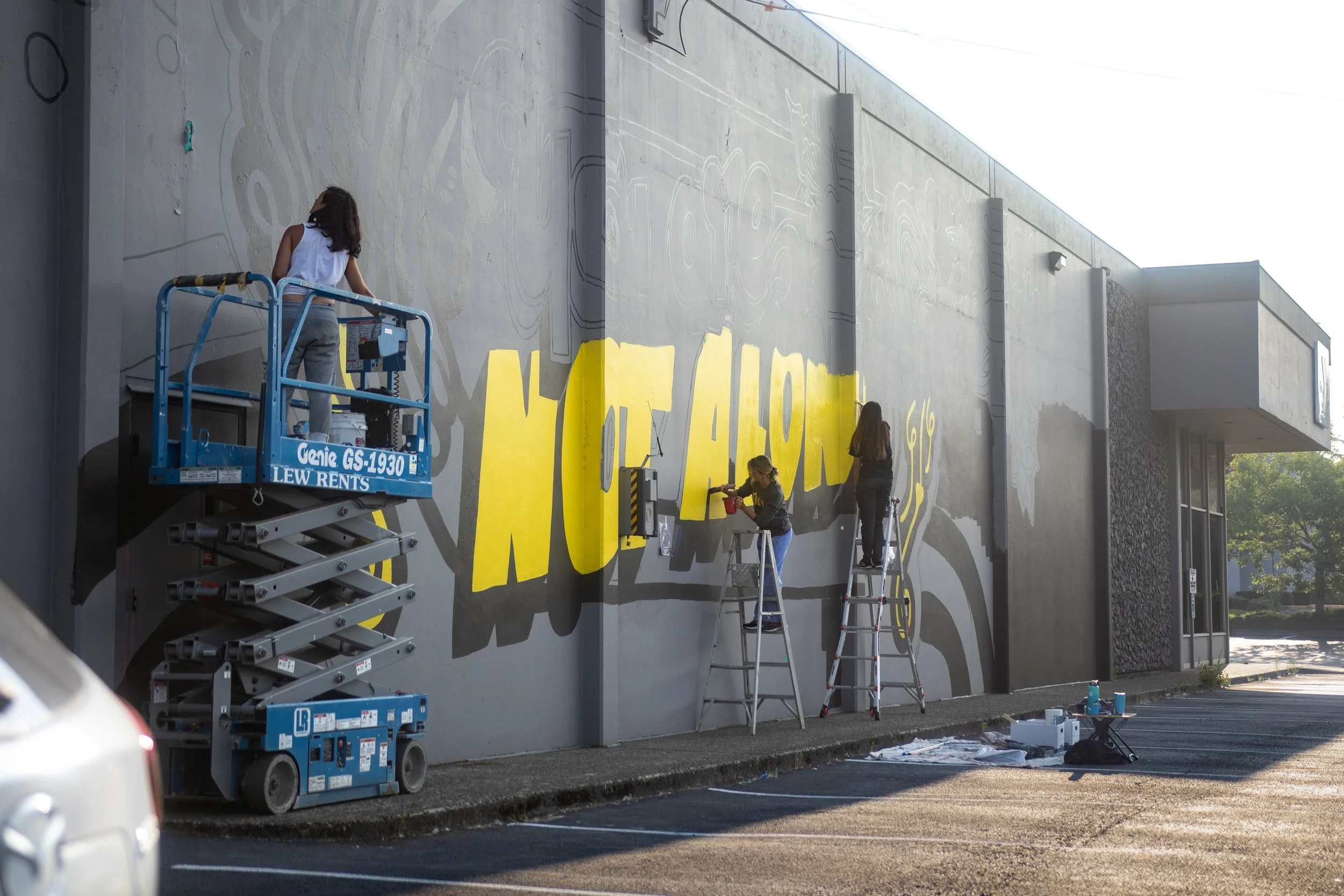 Two artists painting the yellow "You Are Not Alone" mural on a grey wall at the Olympia YMCA, using a scissor lift and a ladder.