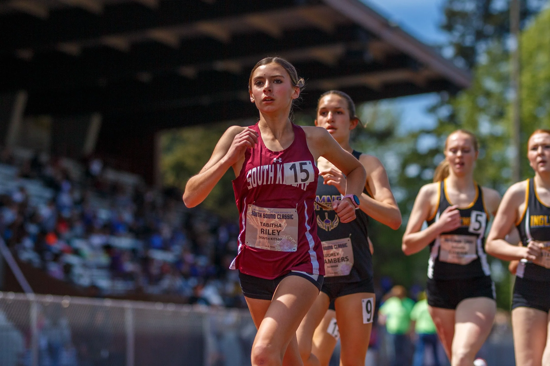 South Kitsap High School athlete Tabitha Riley wearing a maroon uniform and bib number 15, running ahead of three competitors on an outdoor track with stadium seating in the background.