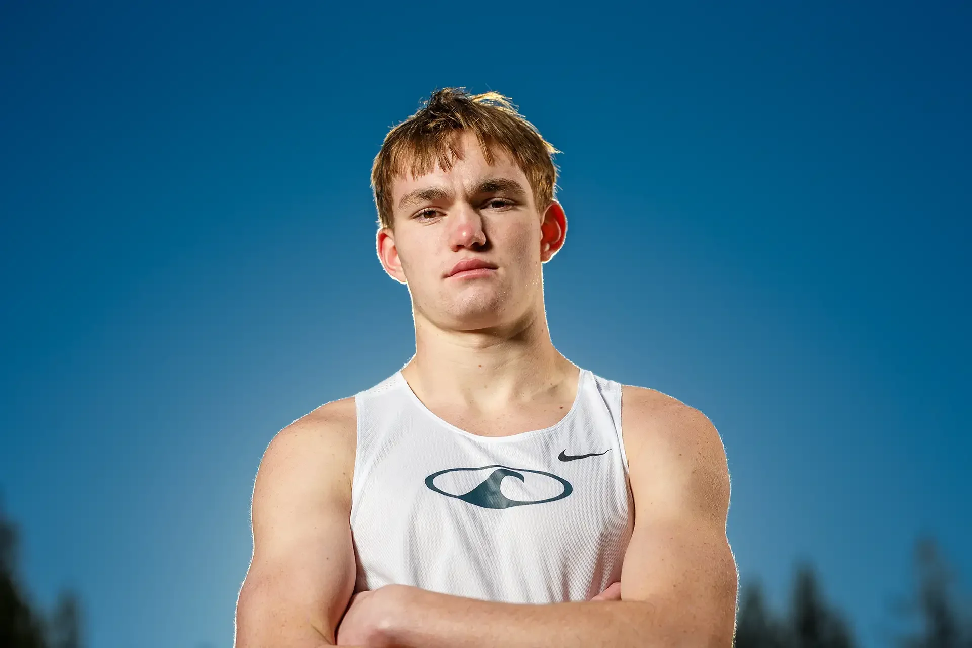 Athletic high school senior boy in a white running singlet with arms crossed looking serious against a deep blue sky at Sehmel Homestead Park.