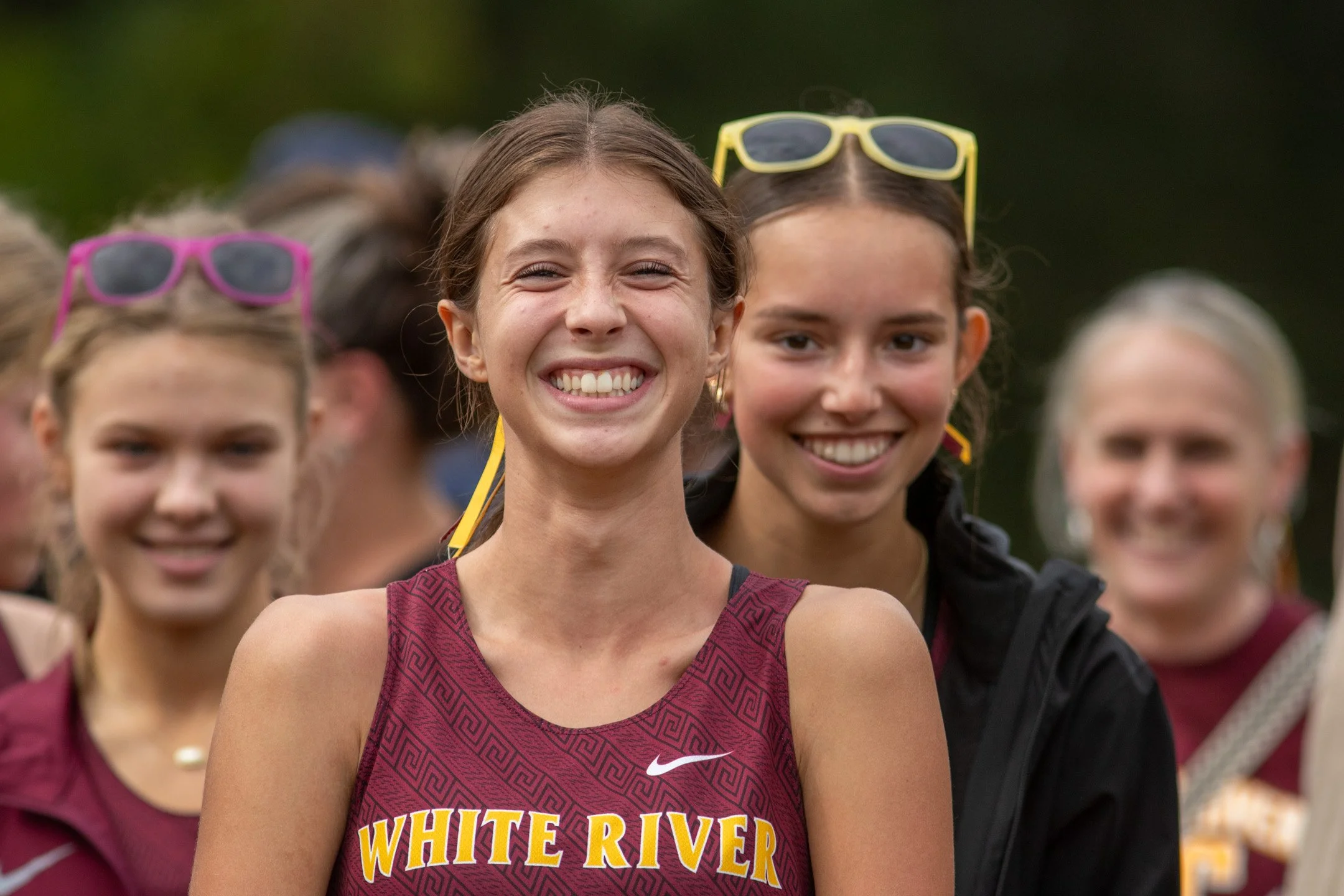 Three White River cross country athletes in maroon jerseys smiling together before a race, one wearing yellow sunglasses pushed up on her head.