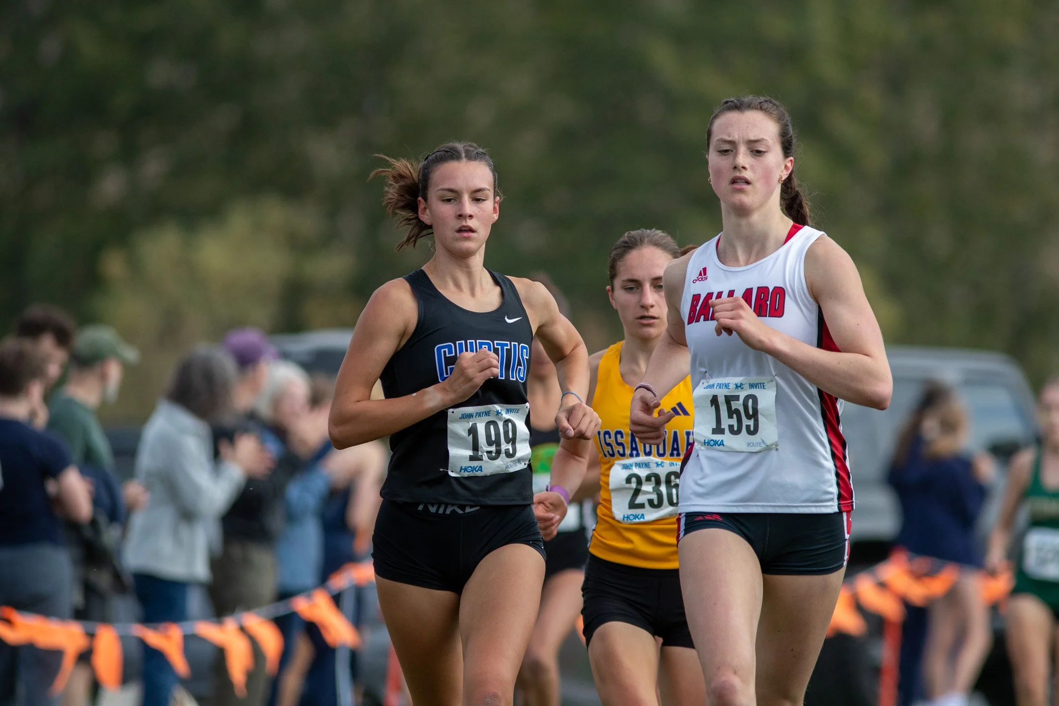 Three high school girls cross country runners—Charlotte White in a black Curtis jersey, Ciana Johnson in a white and red Ballard jersey, and Sally Davis in a gold Issaquah jersey—competing in a tight lead pack along a spectator-lined course.