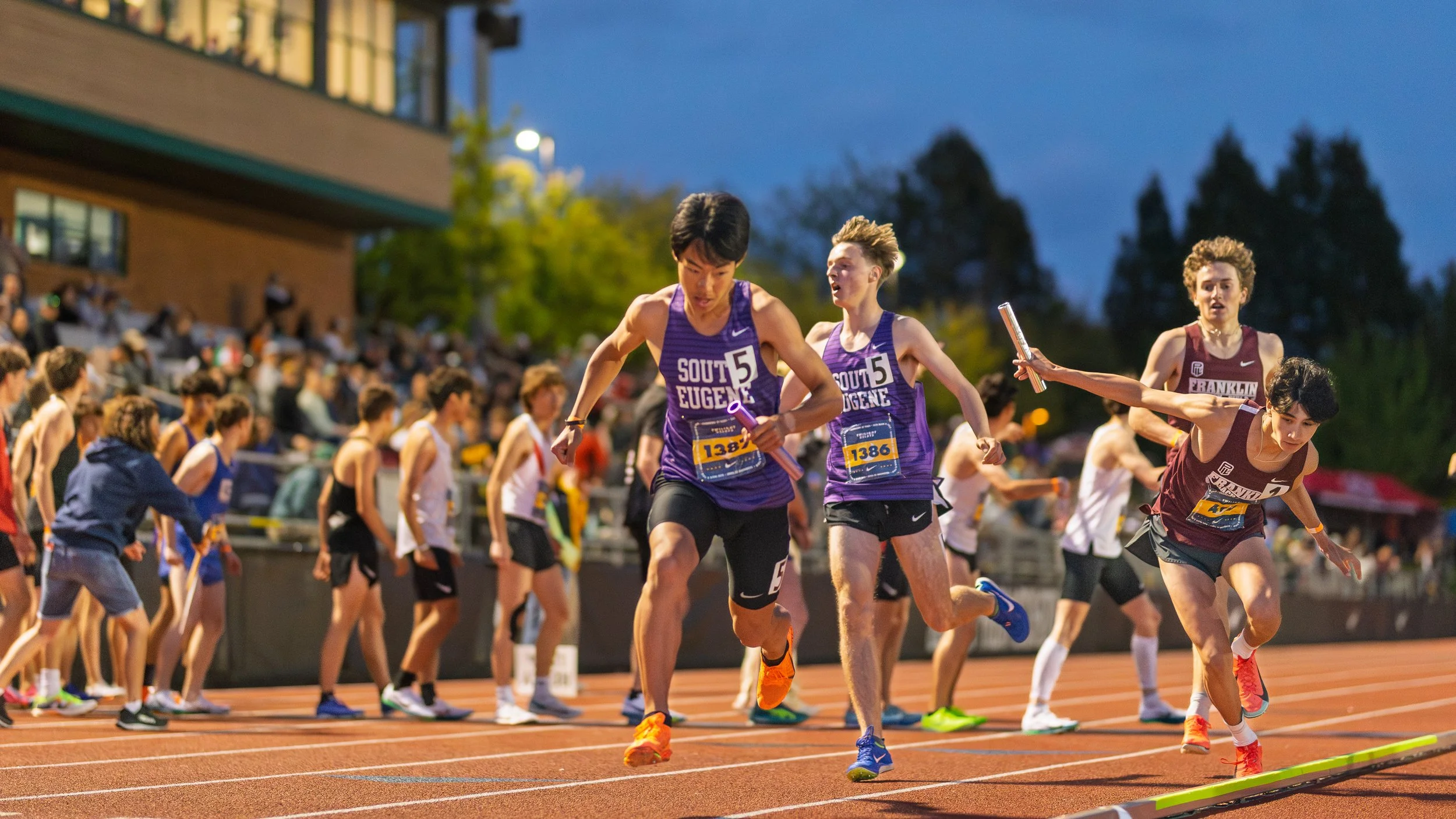 South Eugene athletes wearing purple jerseys race during evening track meet. Franklin High runner lunges forward receiving baton handoff. Spectators fill stadium bleachers under twilight sky.
