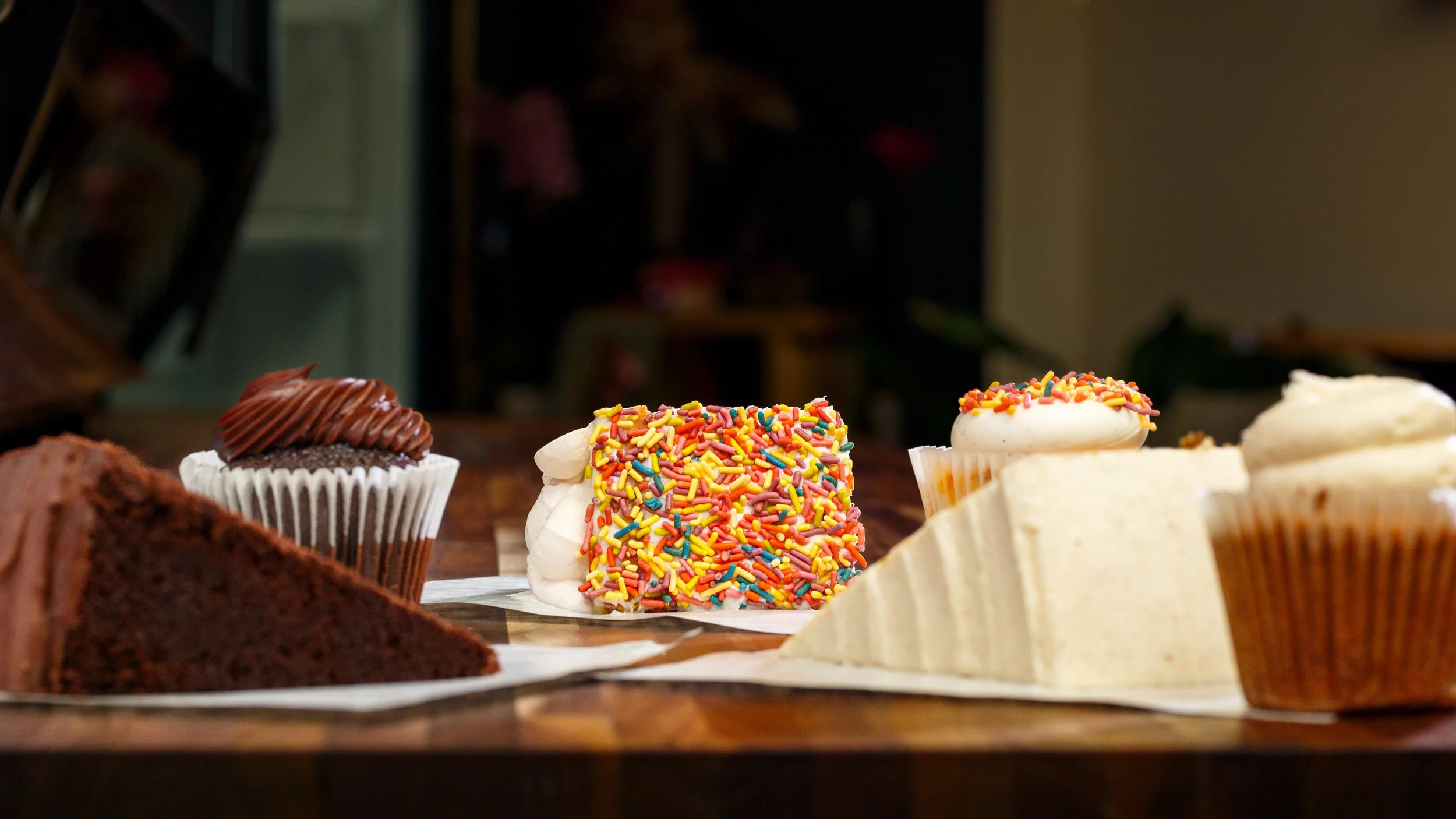 An array of baked goods including a slice of chocolate cake, cupcakes with rich frosting, and a heavily sprinkled treat lined up on a rustic wooden countertop at Tender Velvet Rebel Oven.