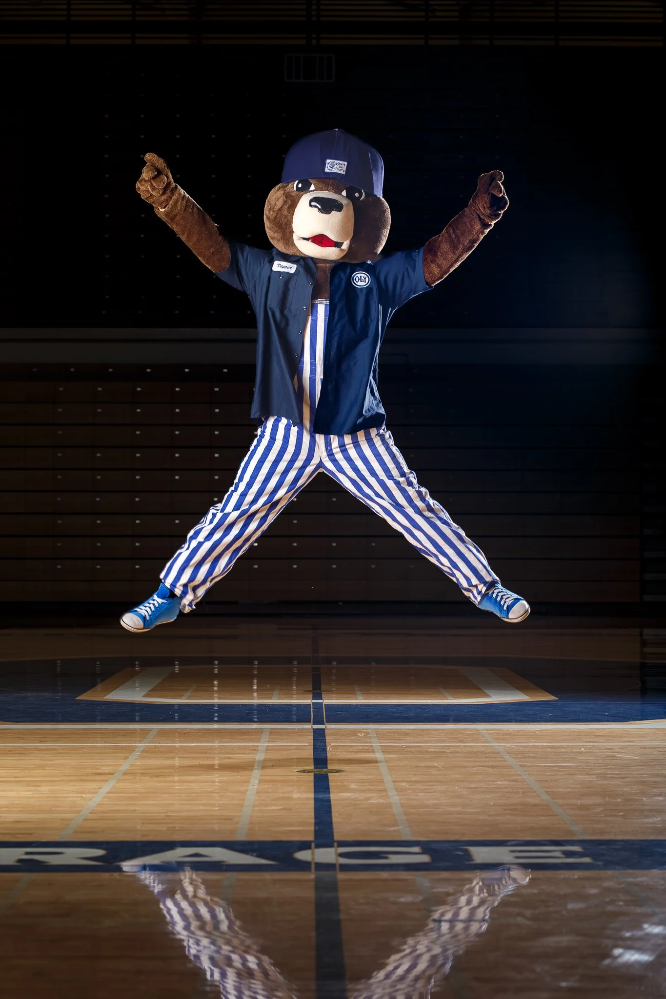 Olympia High School mascot Pepper wearing blue and white striped overalls jumping in the air with arms raised against a dark background.