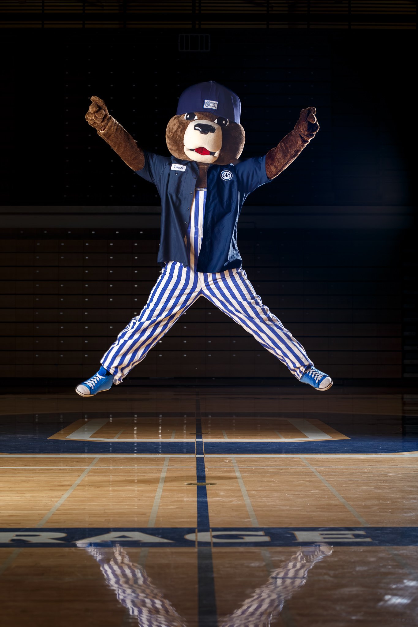 Olympia High School mascot Pepper wearing blue and white striped overalls jumping in the air with arms raised against a dark background.