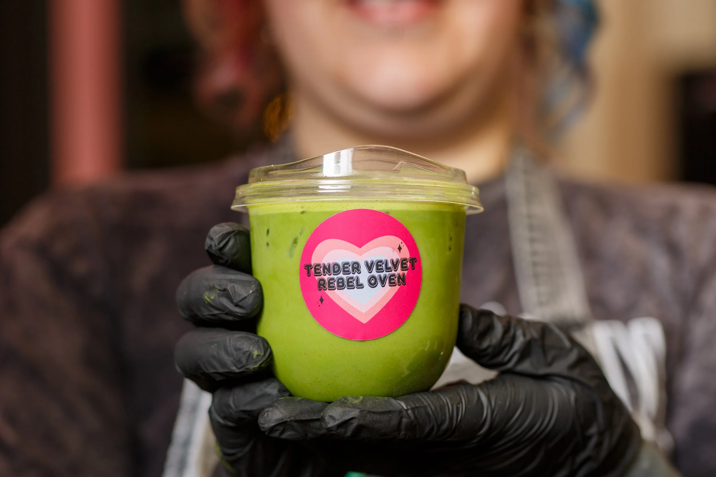 A person wearing black gloves holds a clear plastic cup filled with a bright green matcha drink featuring a pink heart-shaped sticker for Tender Velvet Rebel Oven at 222 Market in Olympia, photographed by Russell Moore.