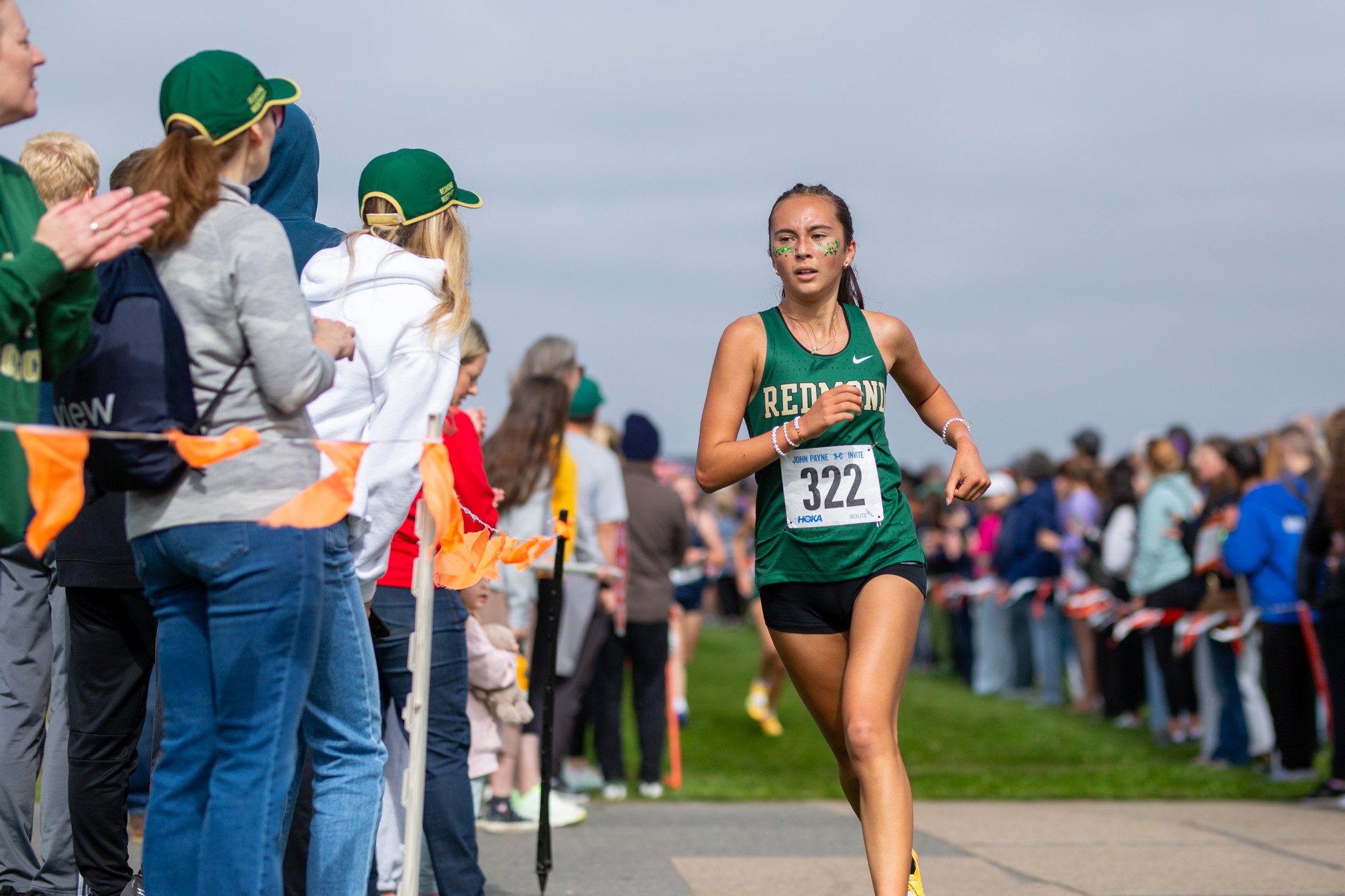 Redmond runner Lenorah McGarty in a dark green jersey with bib 322 running along a course path lined with spectators and orange flags.