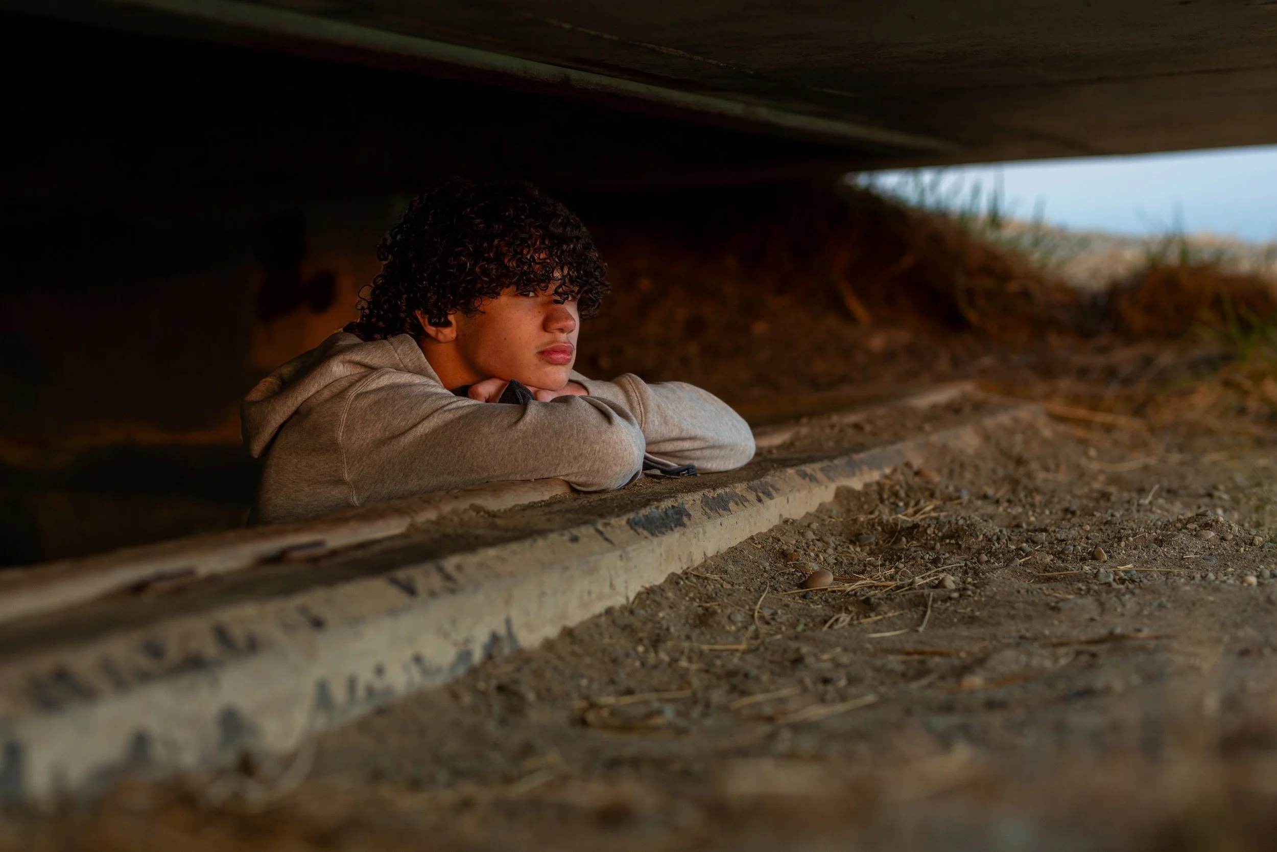 Young man with curly hair resting , leaning on a concrete edge, with dirt and grass in the background.
