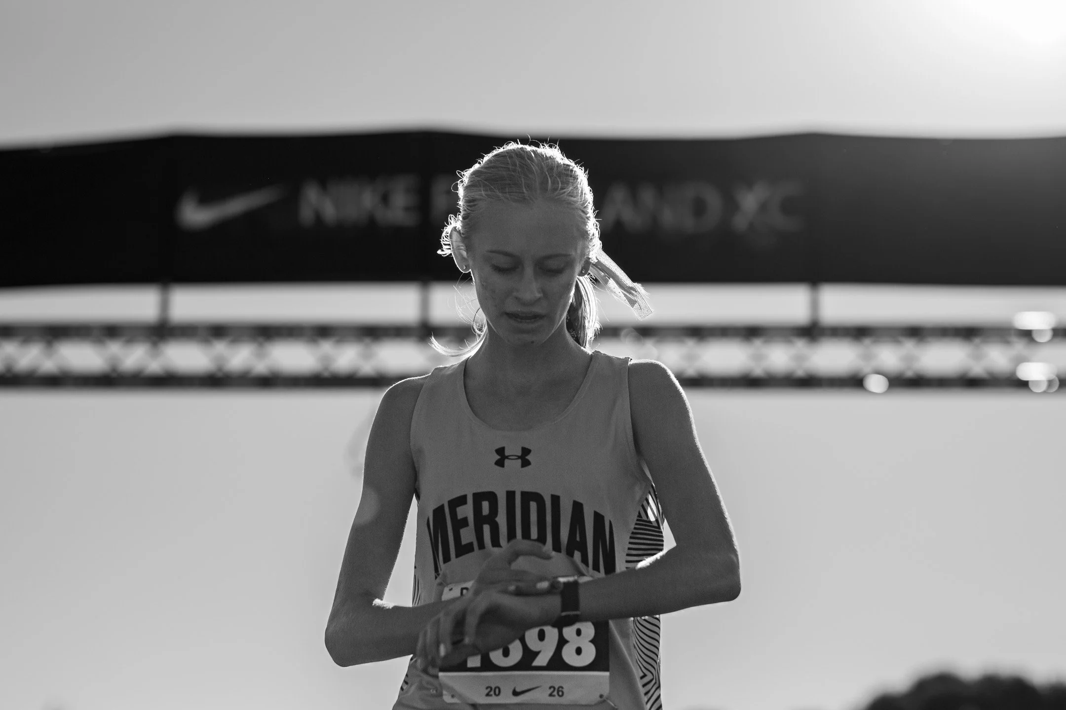 Black-and-white image of Meridian freshman Ridley Strotz looking down at her watch beneath a blurred Nike Portland XC banner.