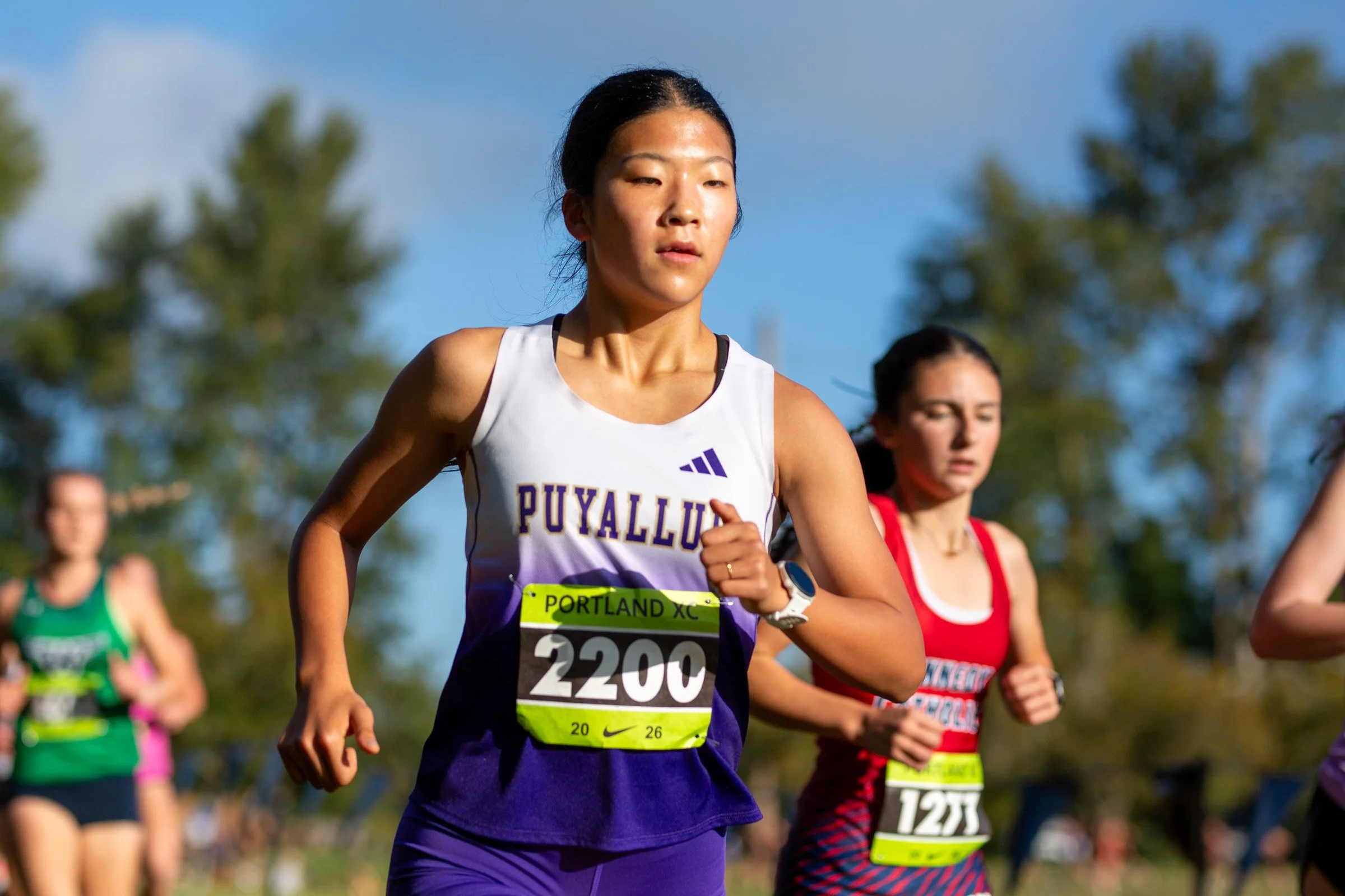 Puyallup High School cross country runner Kyung-Ah Lee in a white and purple Puyallup singlet with bib 2200, running ahead of the pack on a sunny day at the 2026 Nike Portland XC Invitational.