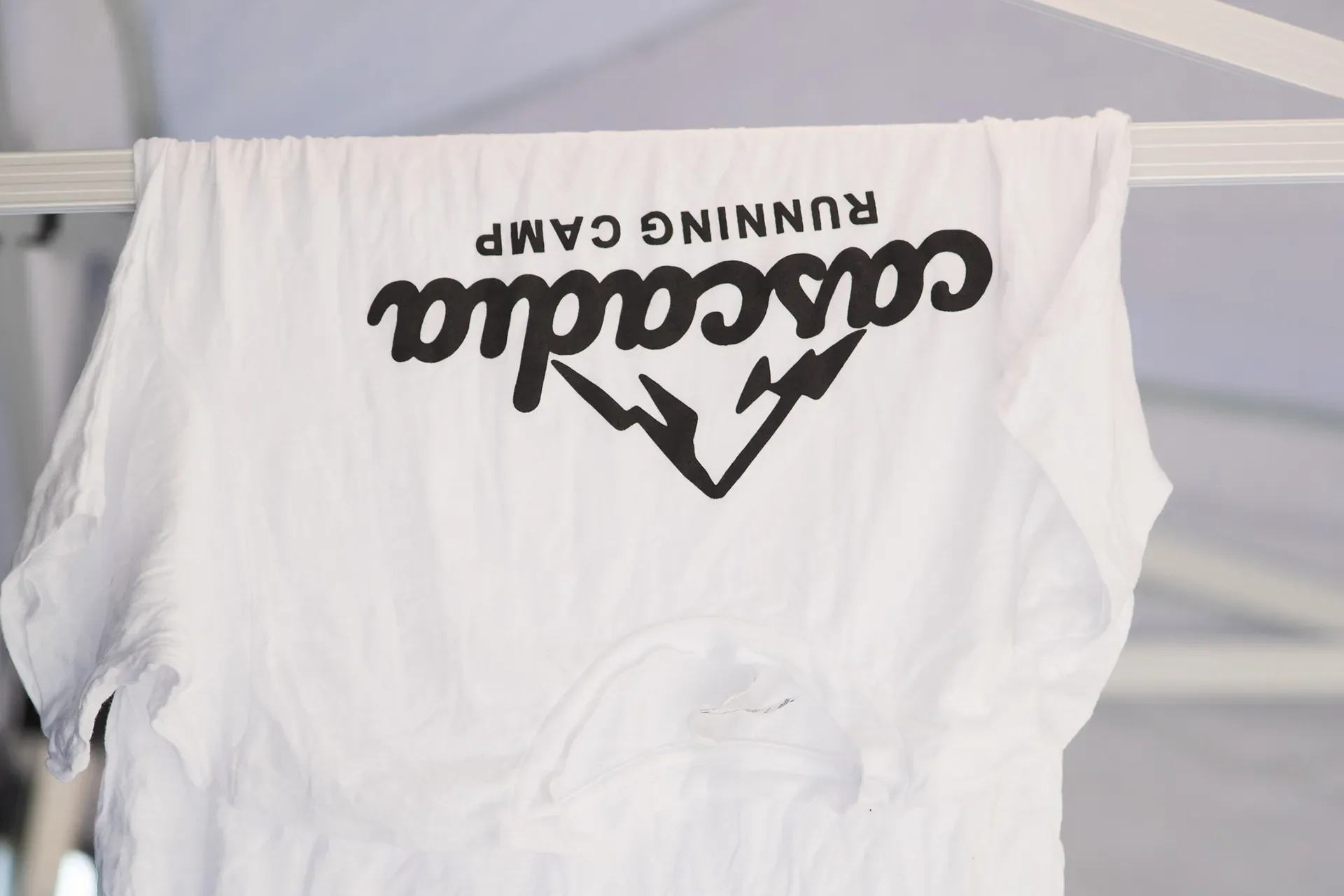 White Cascadia Running Camp t-shirt hanging upside down to dry from a metal tent frame.