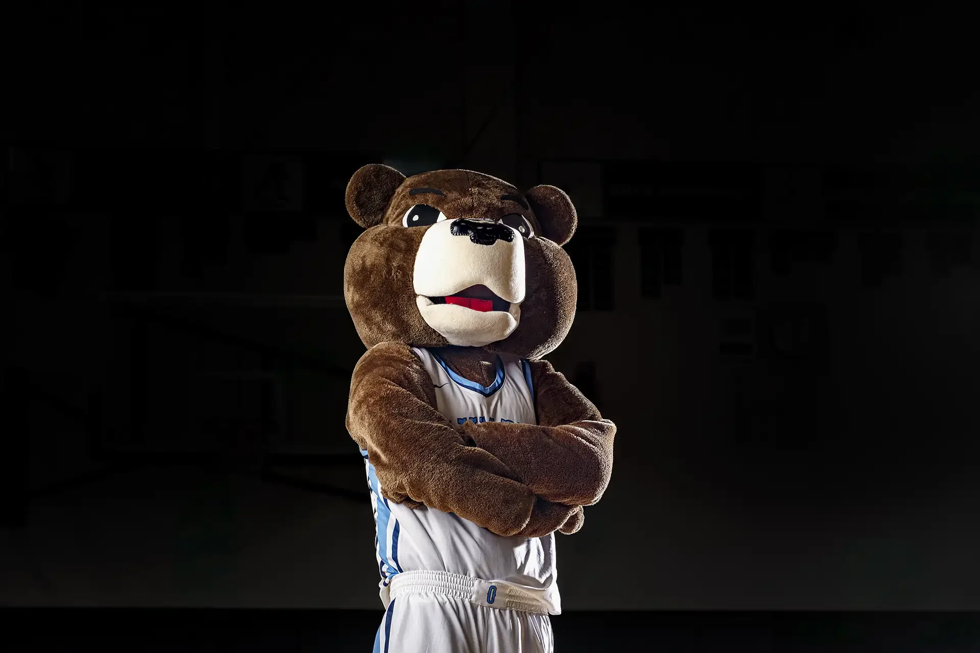 Portrait of Pepper the Bear, the OHS mascot, wearing a white basketball jersey with arms crossed, lit with dramatic high-contrast sports lighting.