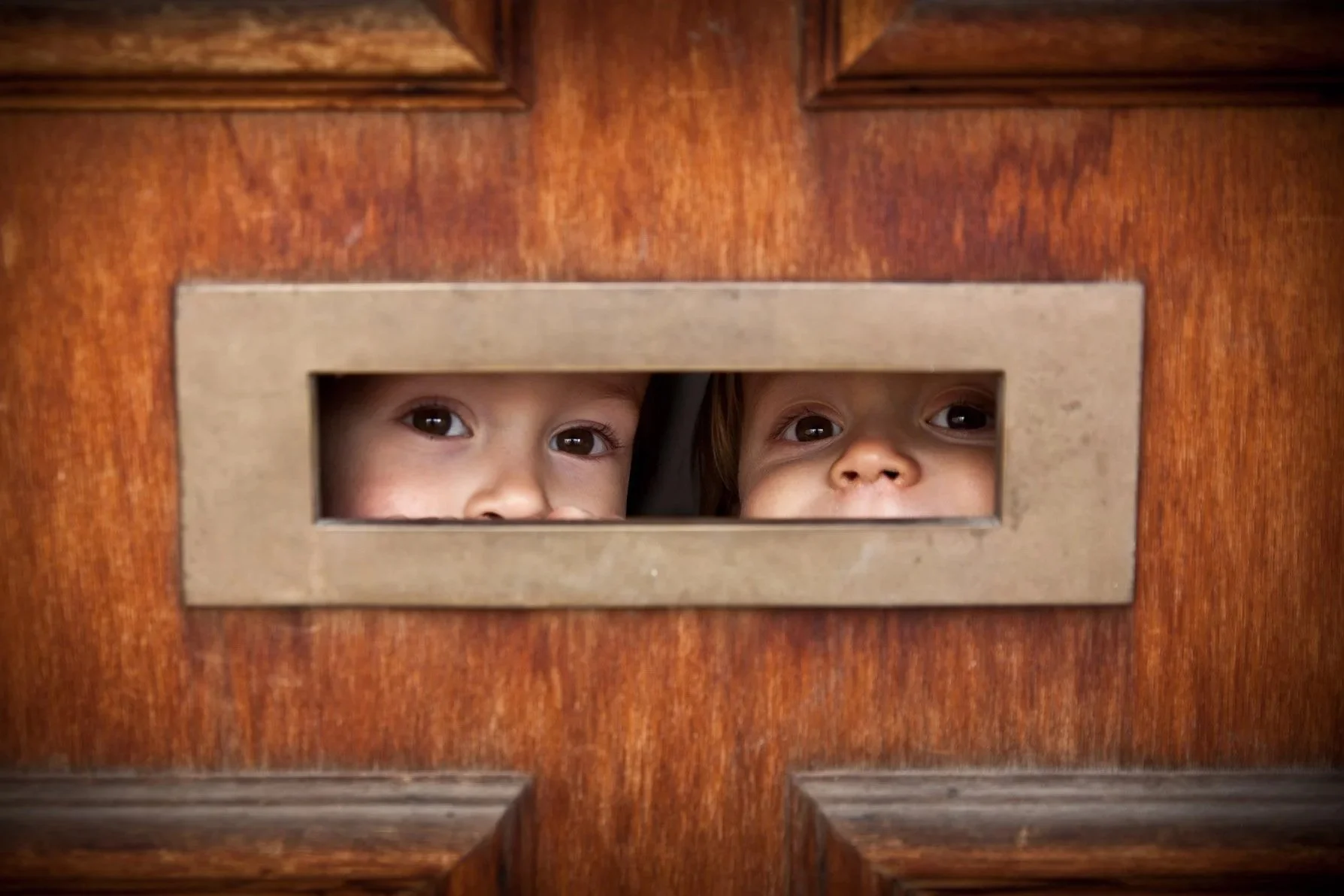 Letterbox, by Melbourne photographer, Ross Calia. Two children looking through a narrow rectangular mail slot in a wooden door.