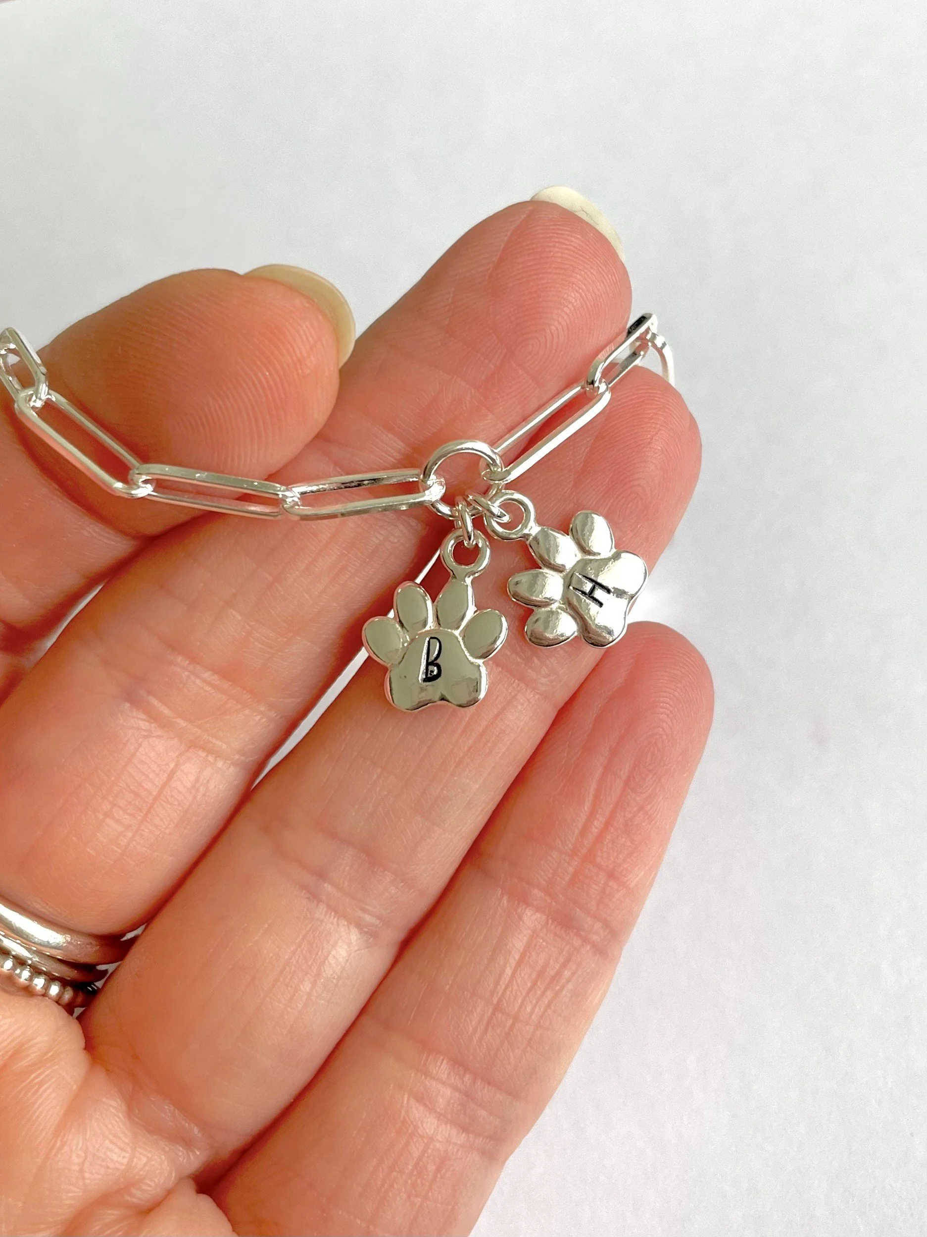 Silver bracelet with two paw print charms, each engraved with a letter, 'B' and 'H', held between fingers against a plain background.