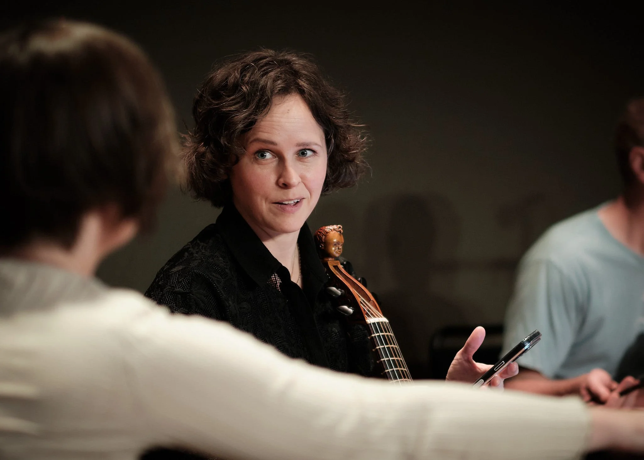 En kvinne med kort, krøllete hår, som ser ut til å snakke, holder en telefon i hånden og har en fiolin med tau og hodehorn på hennes skulder. Hun er omgitt av andre personer som også holder telefoner, i et innendørs rom.