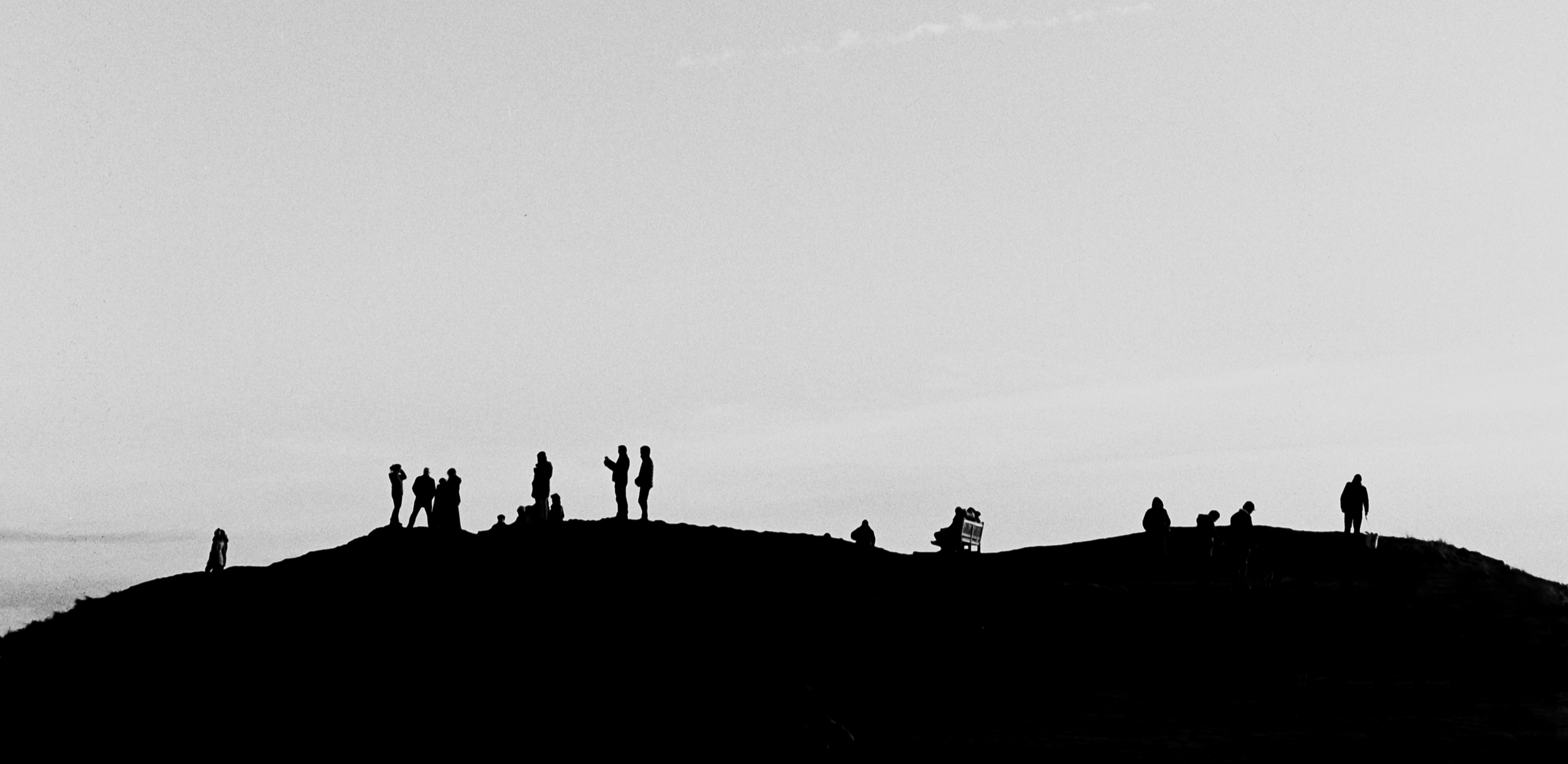 Silhouettes of people standing, sitting, and walking on a hilltop during sunset or sunrise, with a mostly clear sky in the background.
