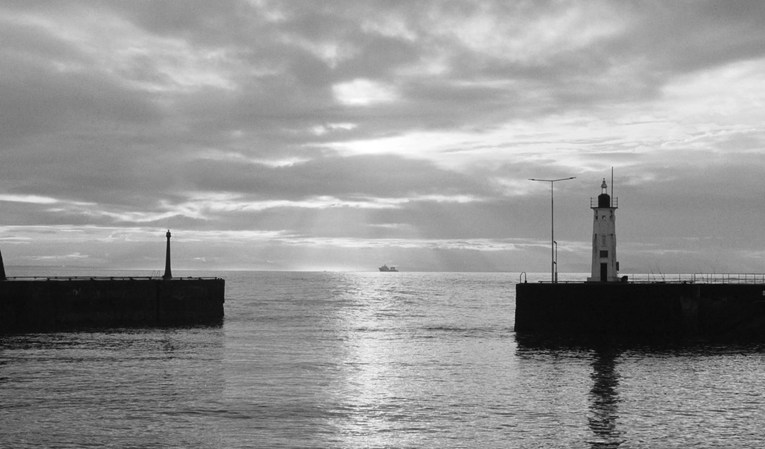 Black and white photo of a harbor with two breakwaters and lighthouses, with a ship in the distance on the water, and cloudy sky above.