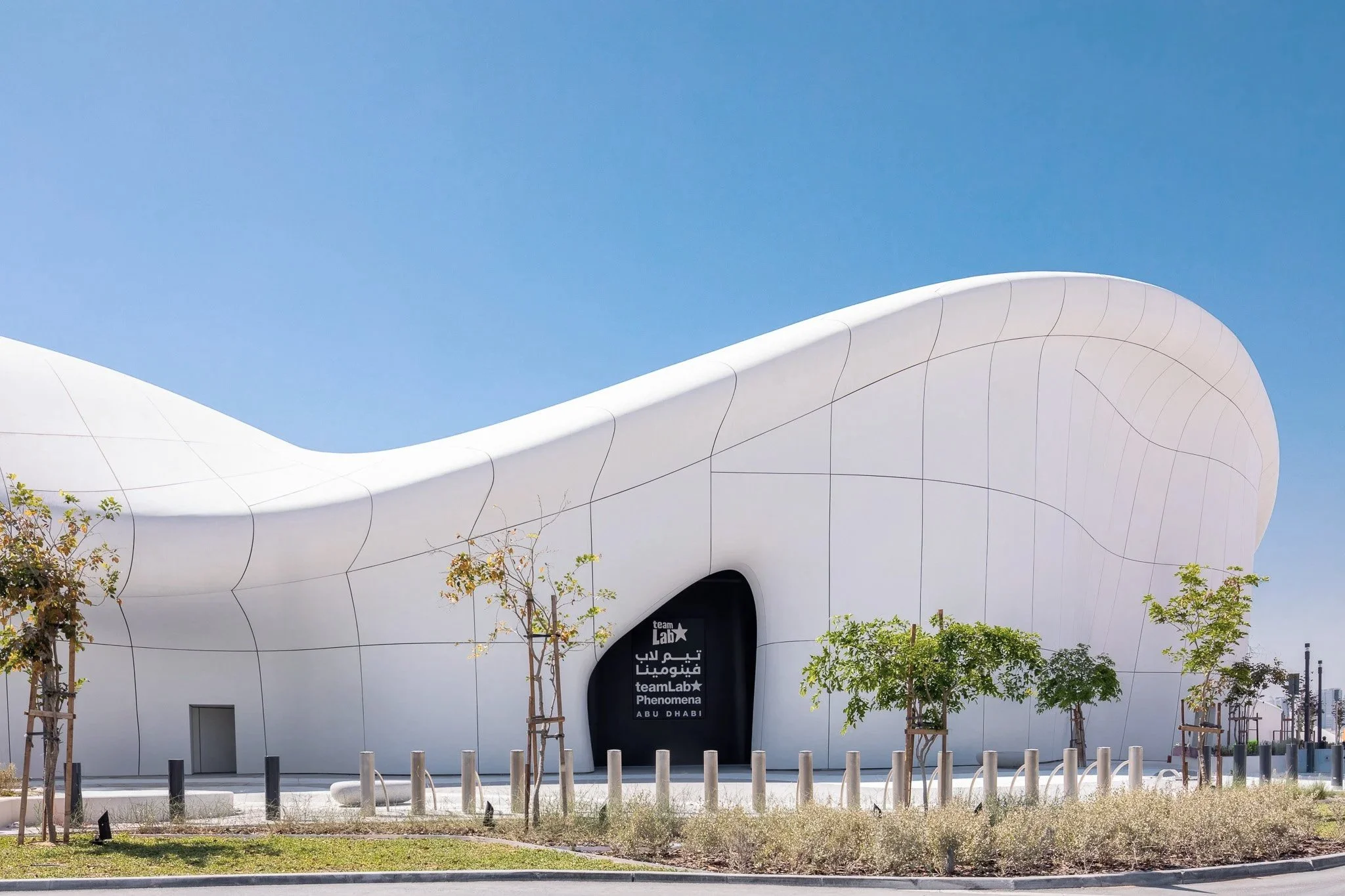 Modern architectural building with smooth white curved exterior, surrounded by young trees and a clear blue sky