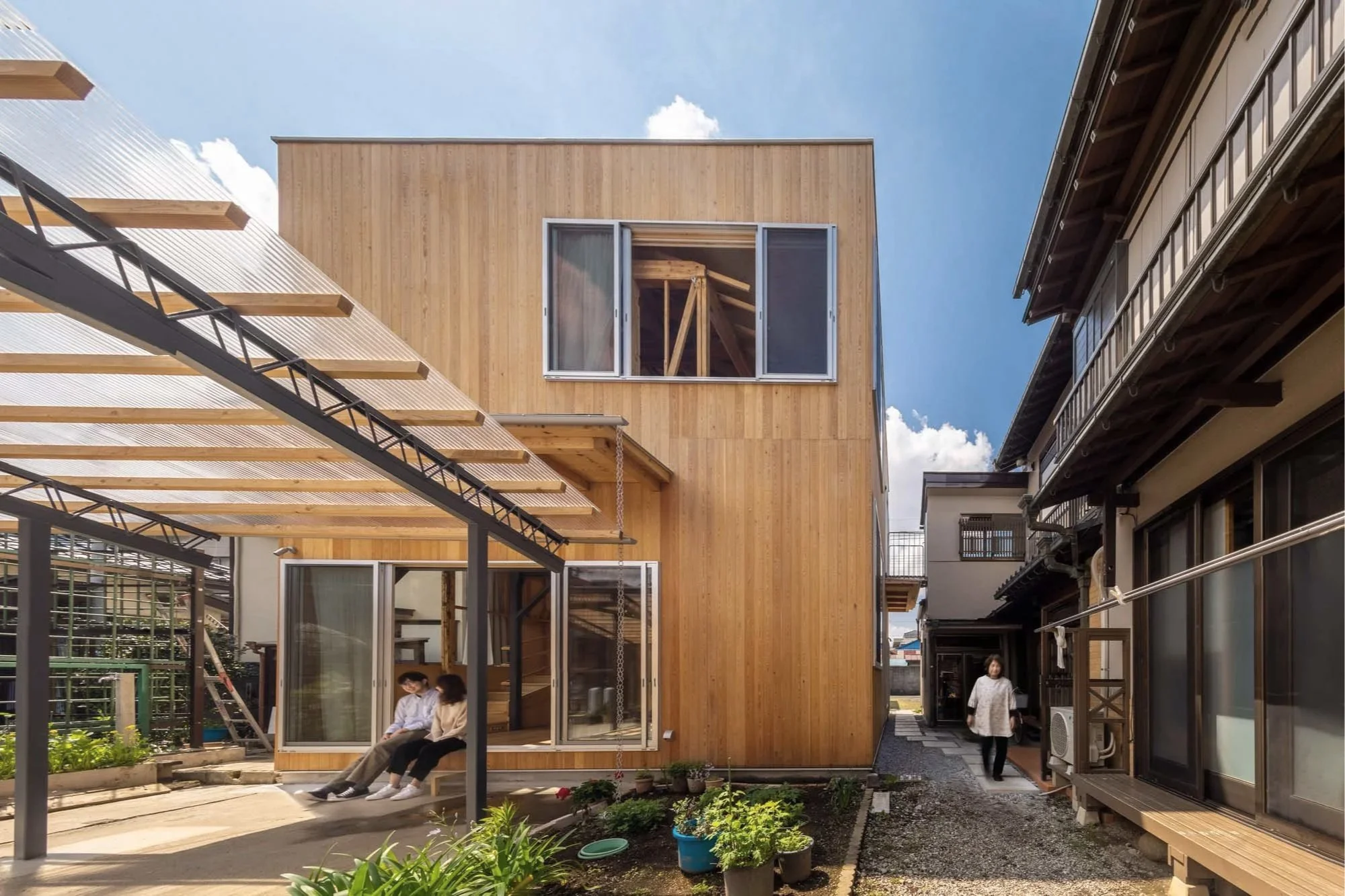 A modern wooden house with open windows, a small garden with potted plants, and two people sitting outside under a metal and plastic canopy, with another person walking along a gravel pathway beside traditional-style houses, under a partly cloudy sky