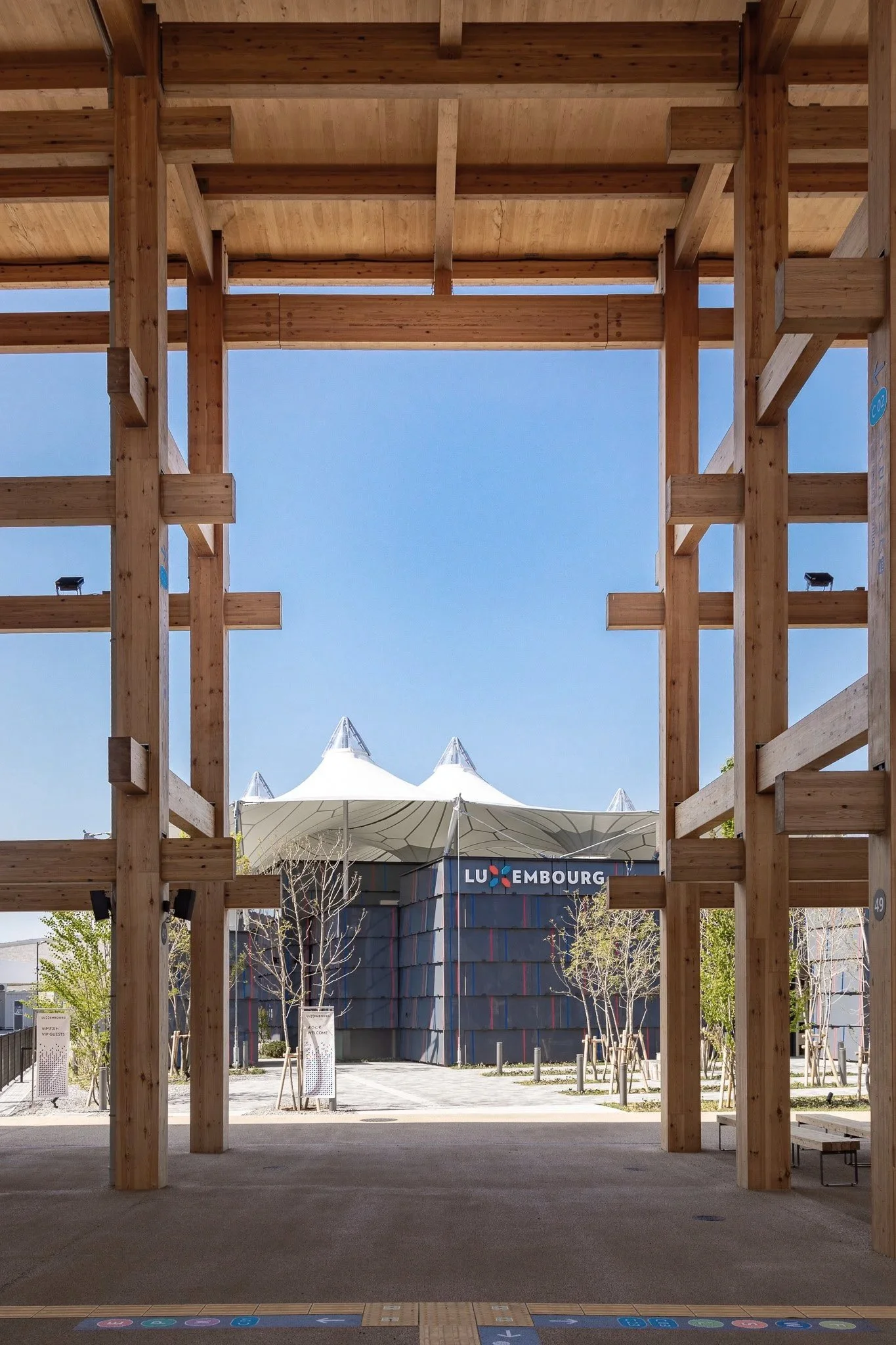 View through a wooden structure towards a modern building with a white tensile fabric roof and a sign that says Luxembourg, under a clear blue sky.