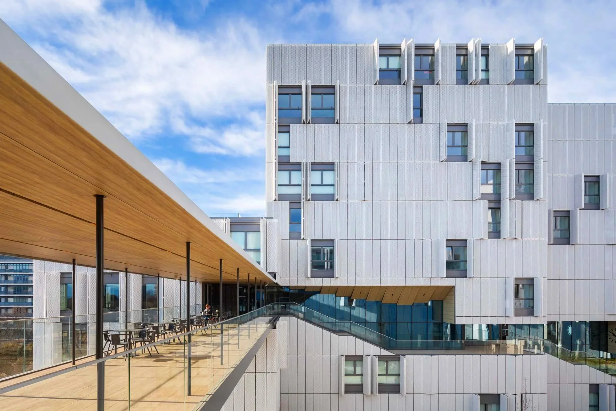 Modern multi-story building with a white exterior, multiple windows, and a rooftop terrace with outdoor seating and a wooden overhang, under a partly cloudy sky.