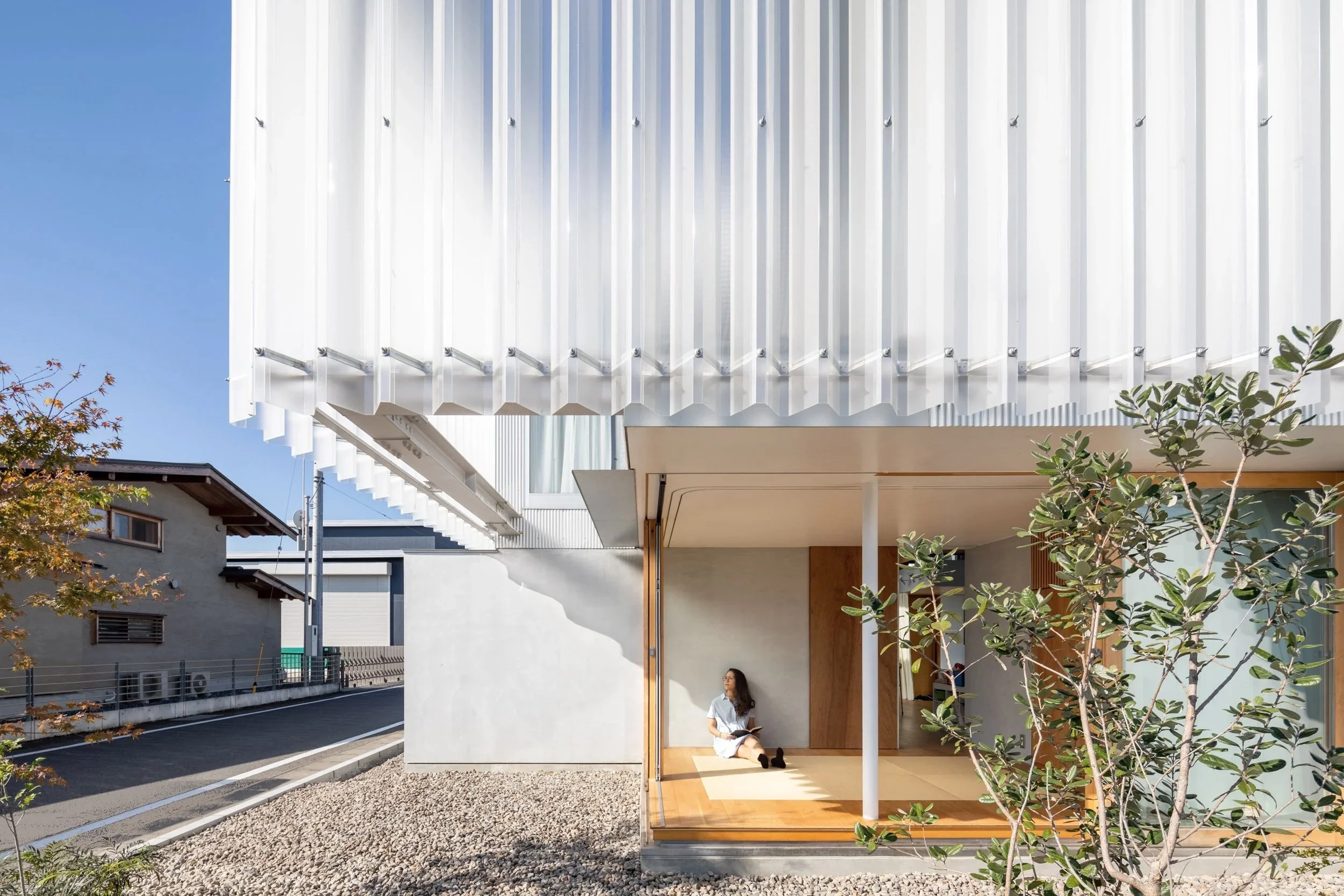 A modern house with a white corrugated metal roof and a woman sitting on the wooden floor of the porch reading a book, with greenery and a neighboring house in the background.
