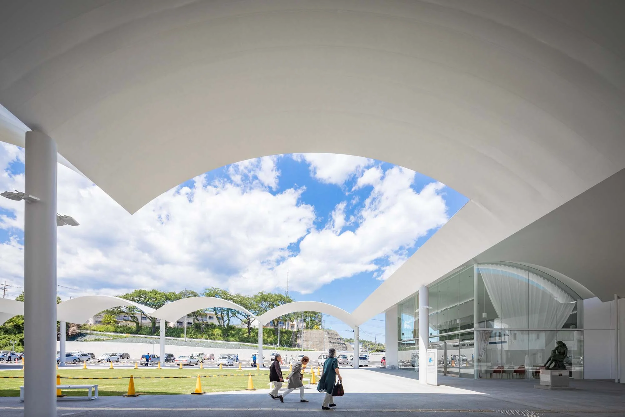 SANAA - Hitachi City Hall - People walk under modern, white, arched structures; blue sky and parked cars background.