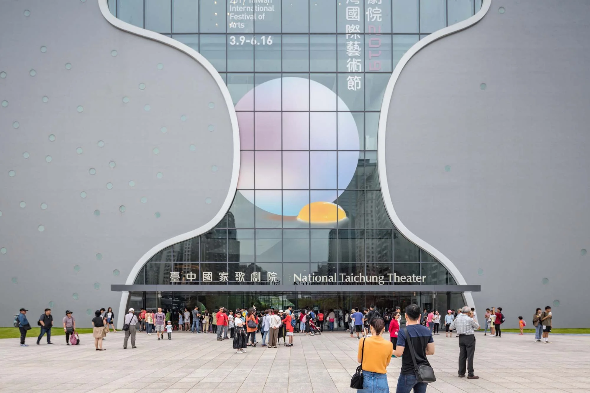 Crowd of people gathered in front of the National Taichung Theater, a modern building with a distinctive curved architectural design and large glass windows.