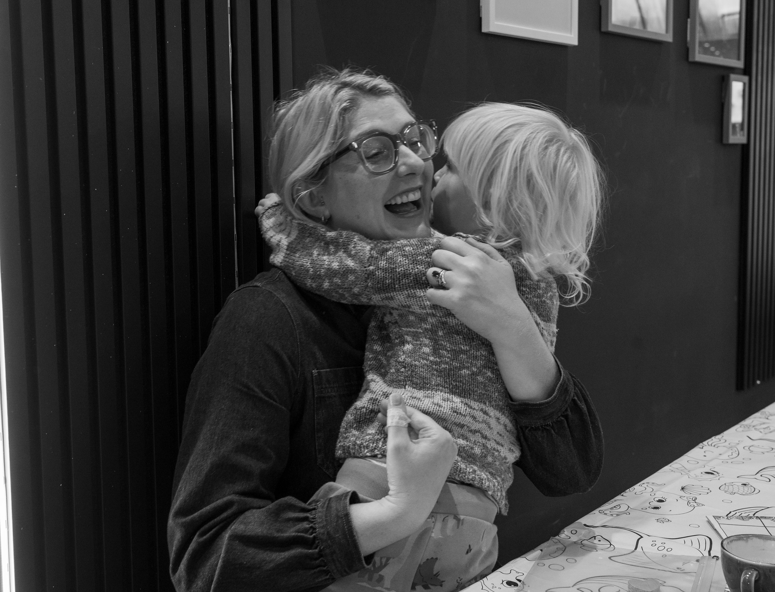 A woman with glasses hugging a young girl who is kissing her on the cheek, both smiling, in a room with a dark wall and framed pictures.